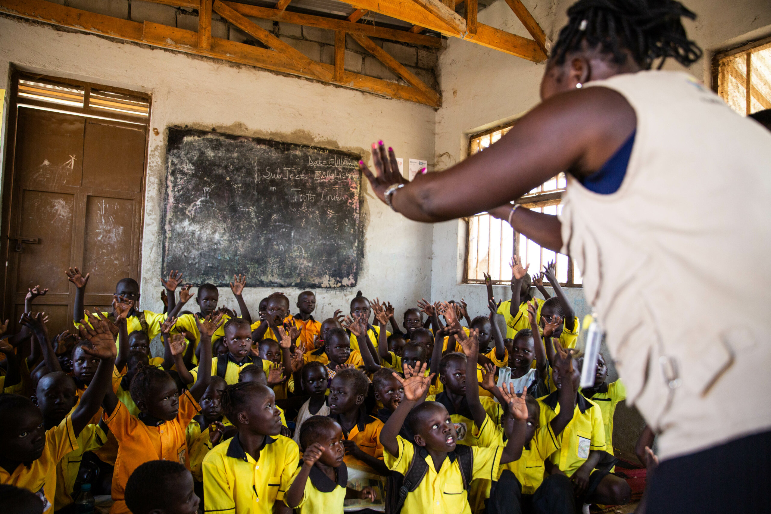 A classroom of learners sitting together on a carpet on the floor of a community nursery class in an IDP camp in Juba, South Sudan, whilst their teacher stands in front of them, signing. The teacher has her back to the camera and raises both her hands, which the children are copying her sign language.