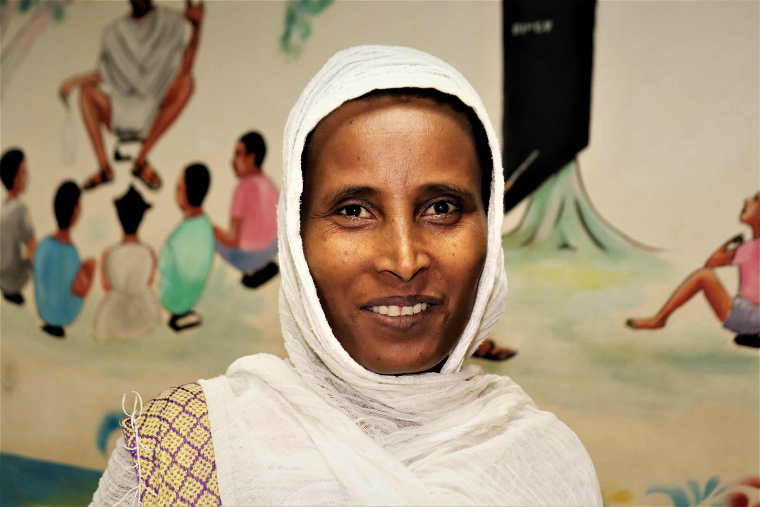 A woman is smiling, wearing a white headdress, after receiving glaucoma treatment. She stands in front of a wall, with drawings of children.