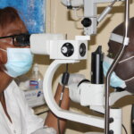 A female doctor, wearing a white jacket, glasses and mask is carrying out an eye exam on a male patient, as part of glaucoma treatment.