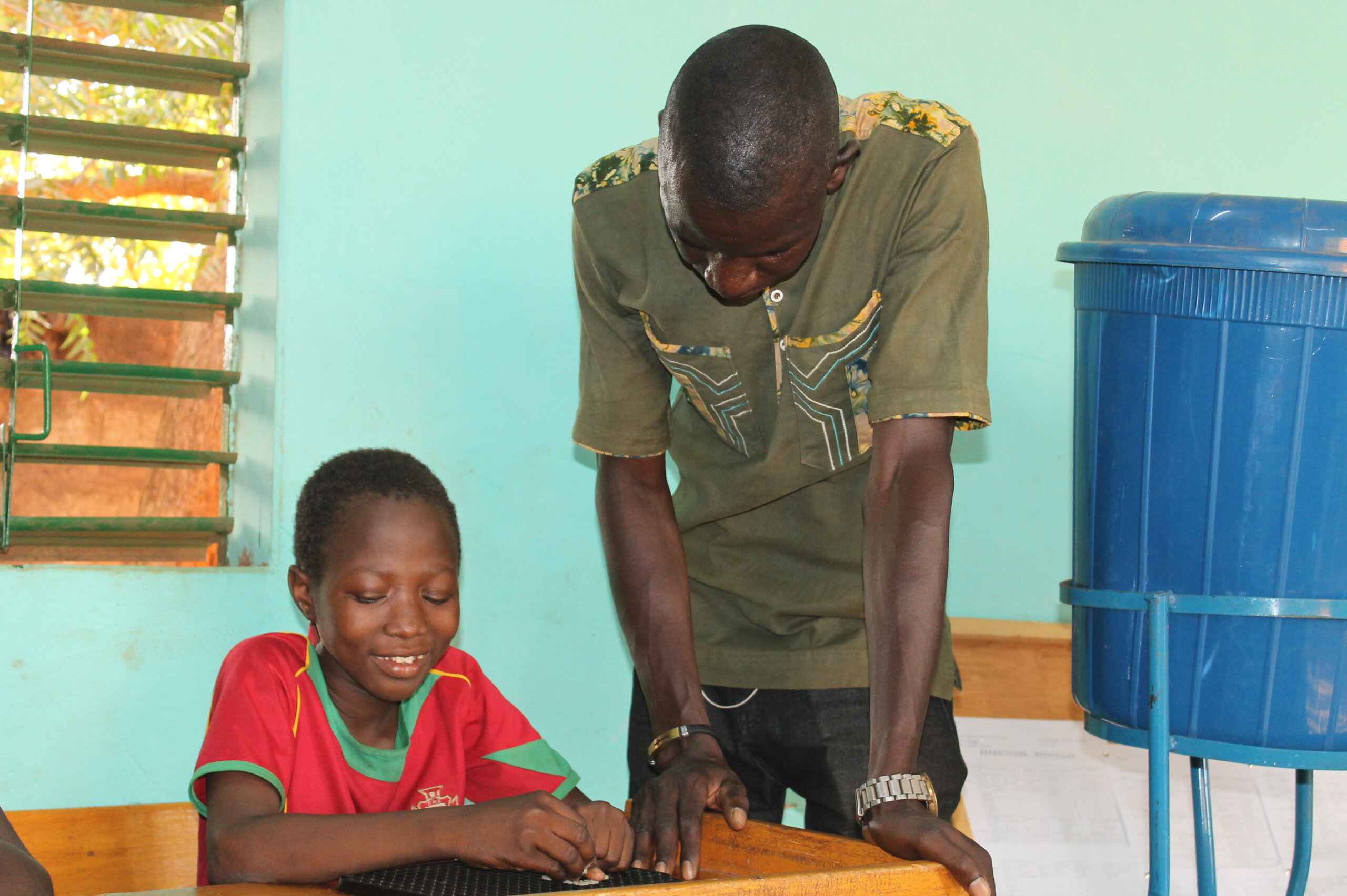 A school girls is sitting at a desk, wearing a red shirt, after she received glaucoma treatment. Her teacher stands next to her, wearing a dark green t-shirt. 