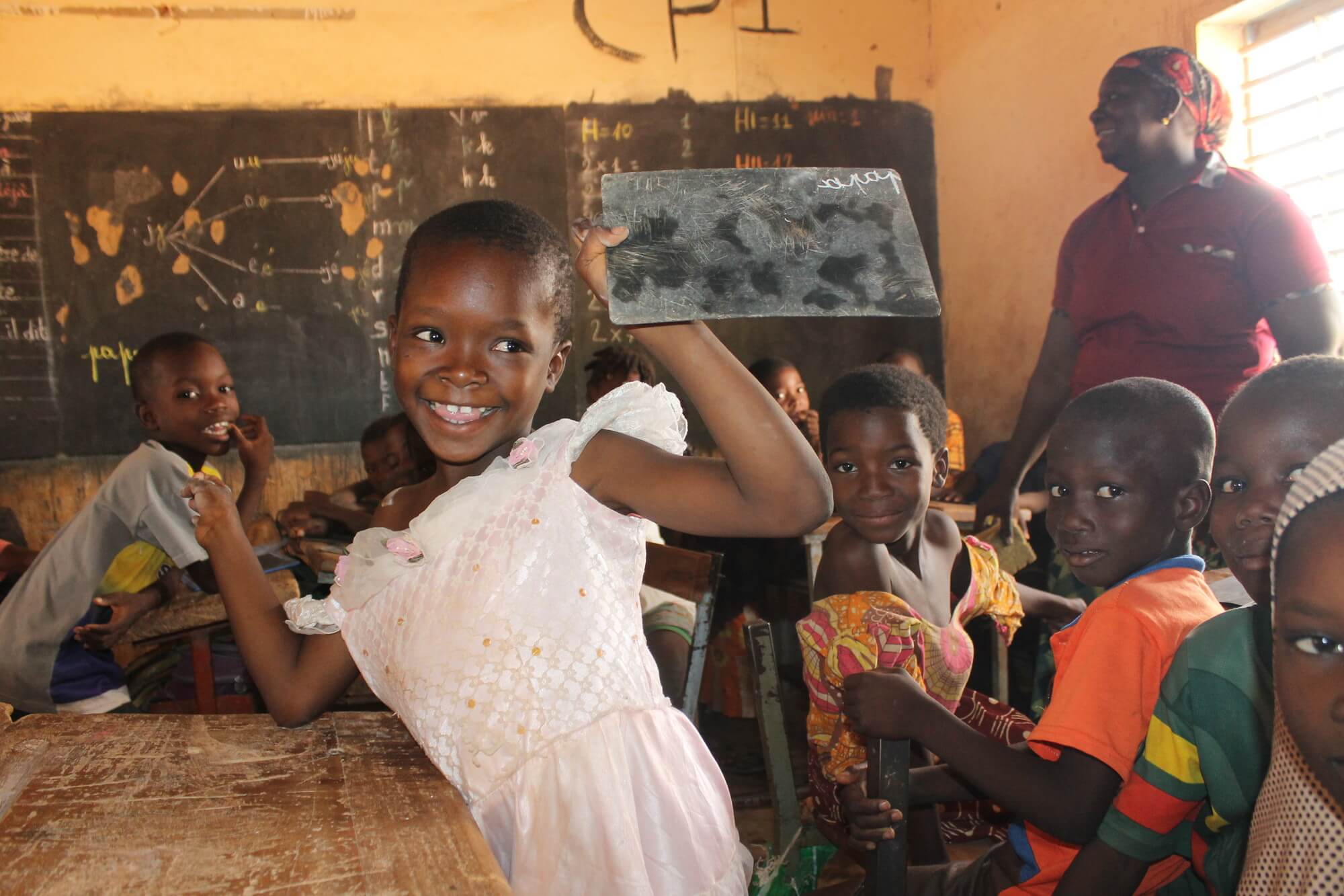 Image of Adissa Kabore, a young girl who has hemiplegia, in a classroom. She is surrounded by classmates and proudly holding a small chalkboard. Adissa received rehabilitation from OCADES (CARITAS), a project partner of Light for the World working on inclusive education in Burkina Faso. She now attends primary school, where her rehabilitation is supported by teachers. Copyright for the photo is Light for the World