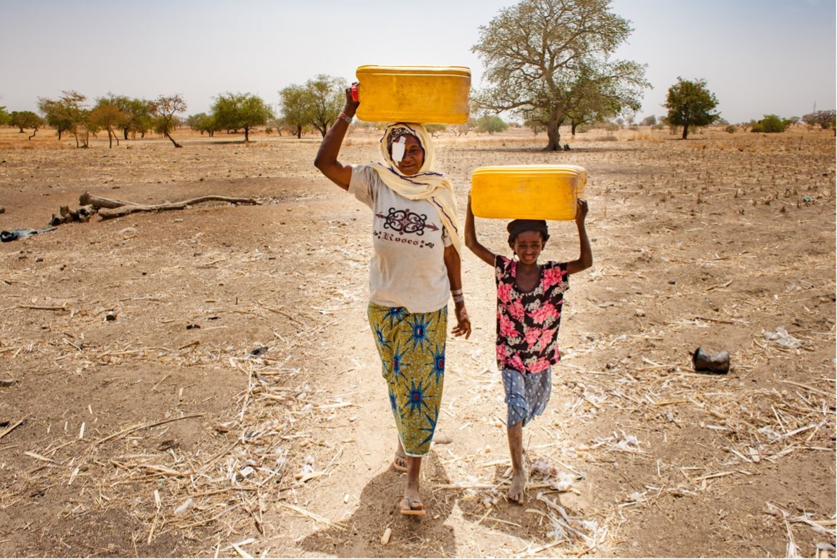 A woman and a girl walk through a field, carrying buckets of water on their heads. Access to clean water and sanitation is critical to preventing the spread of eye infections, which is sustainable development goal 6.