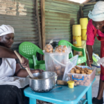 Lylian (left) is wearing a white cap and apron and stirring dough in a bowl, while her colleague (right) is wearing a red dress and apron and handles doughnuts. They are surrounded by baked goods.