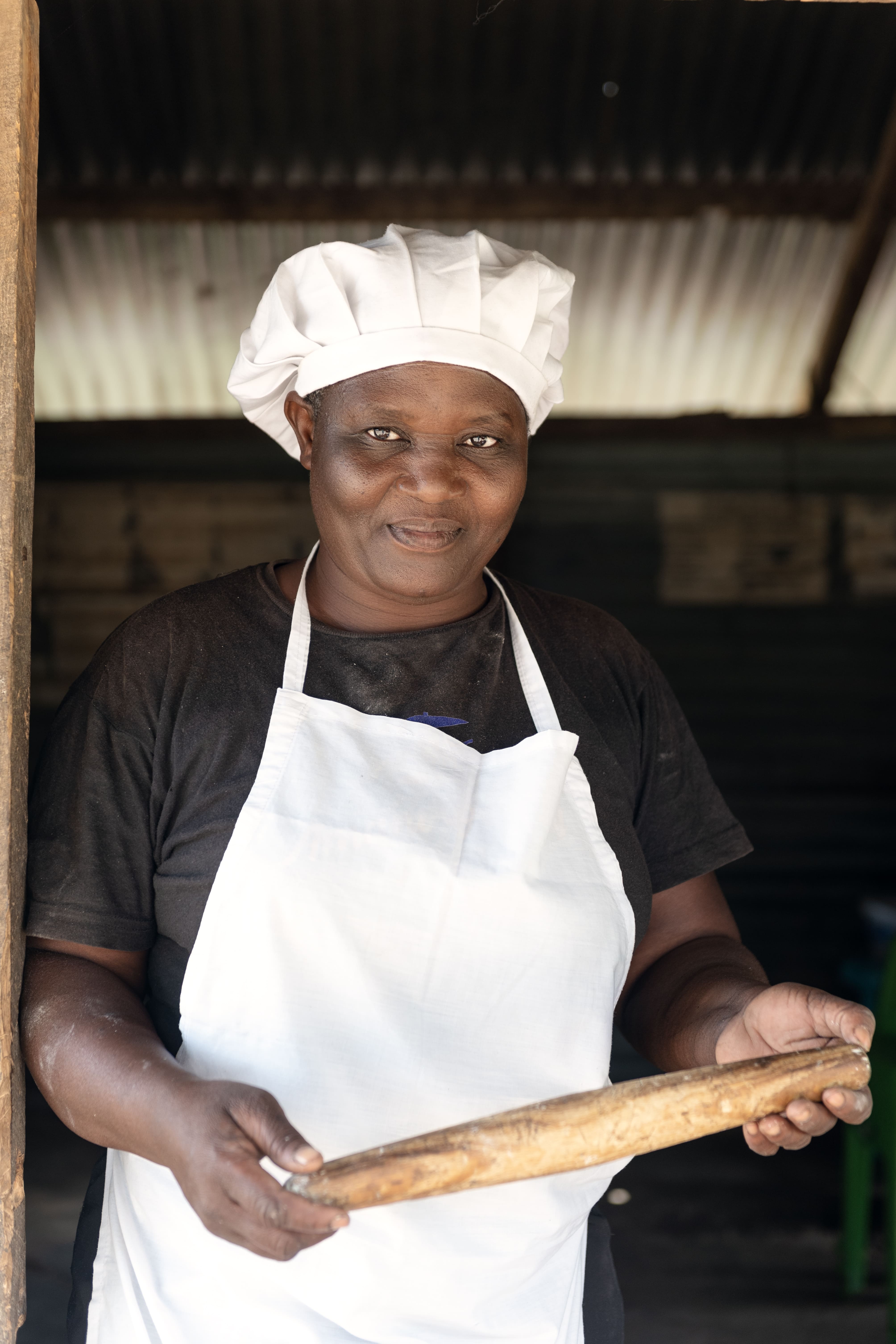 Lylian stands in her doorway, wearing an apron and a white cap, holding a loaf of bread. She is Breaking barriers to economic independence for people with disabilities.