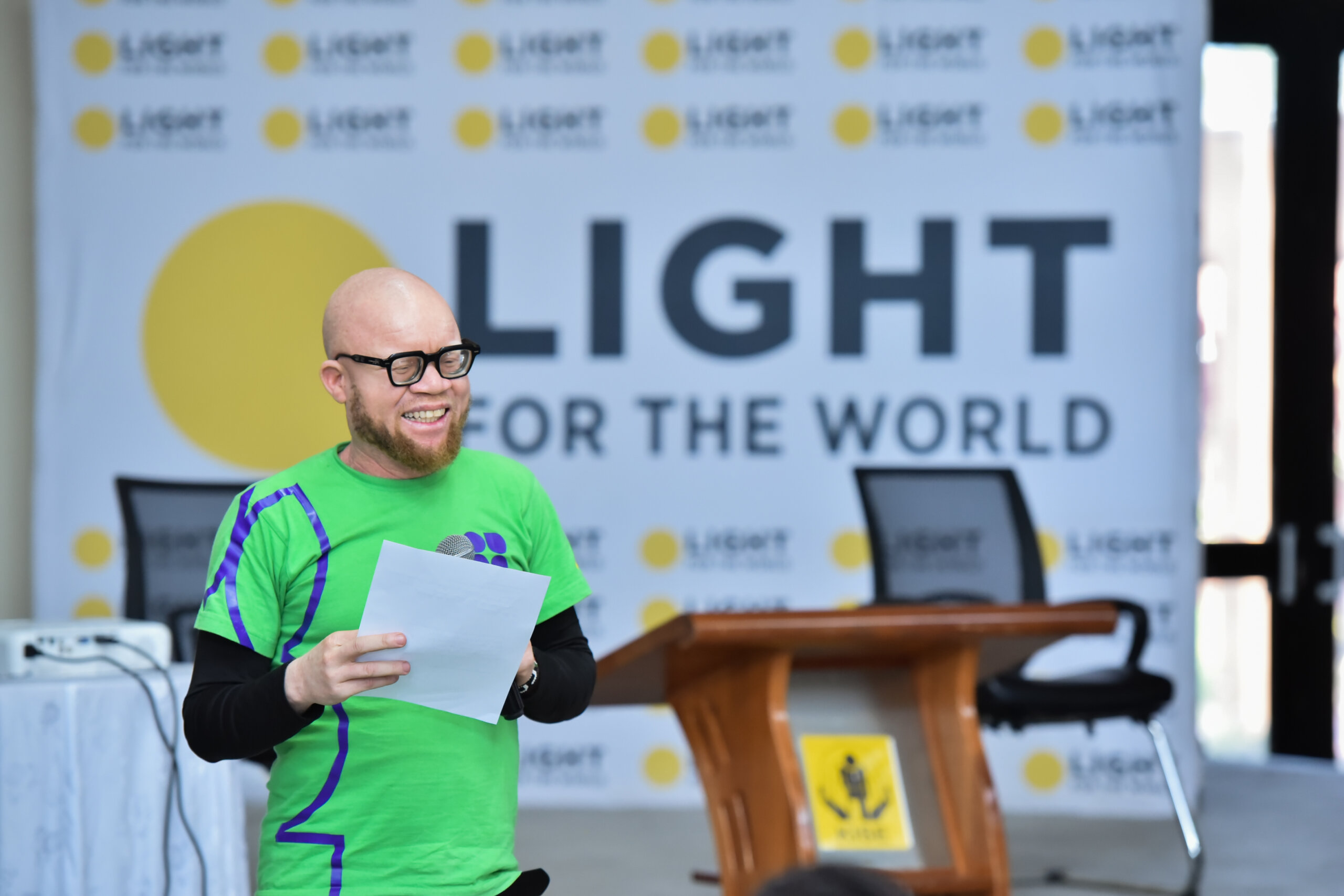Disability rights activist, Collins Ombajo stands on stage, wearing a green top and holding a piece of paper. He is standing in front of a Light for the World sign and breaking barriers to economic independence for people with disabilities.