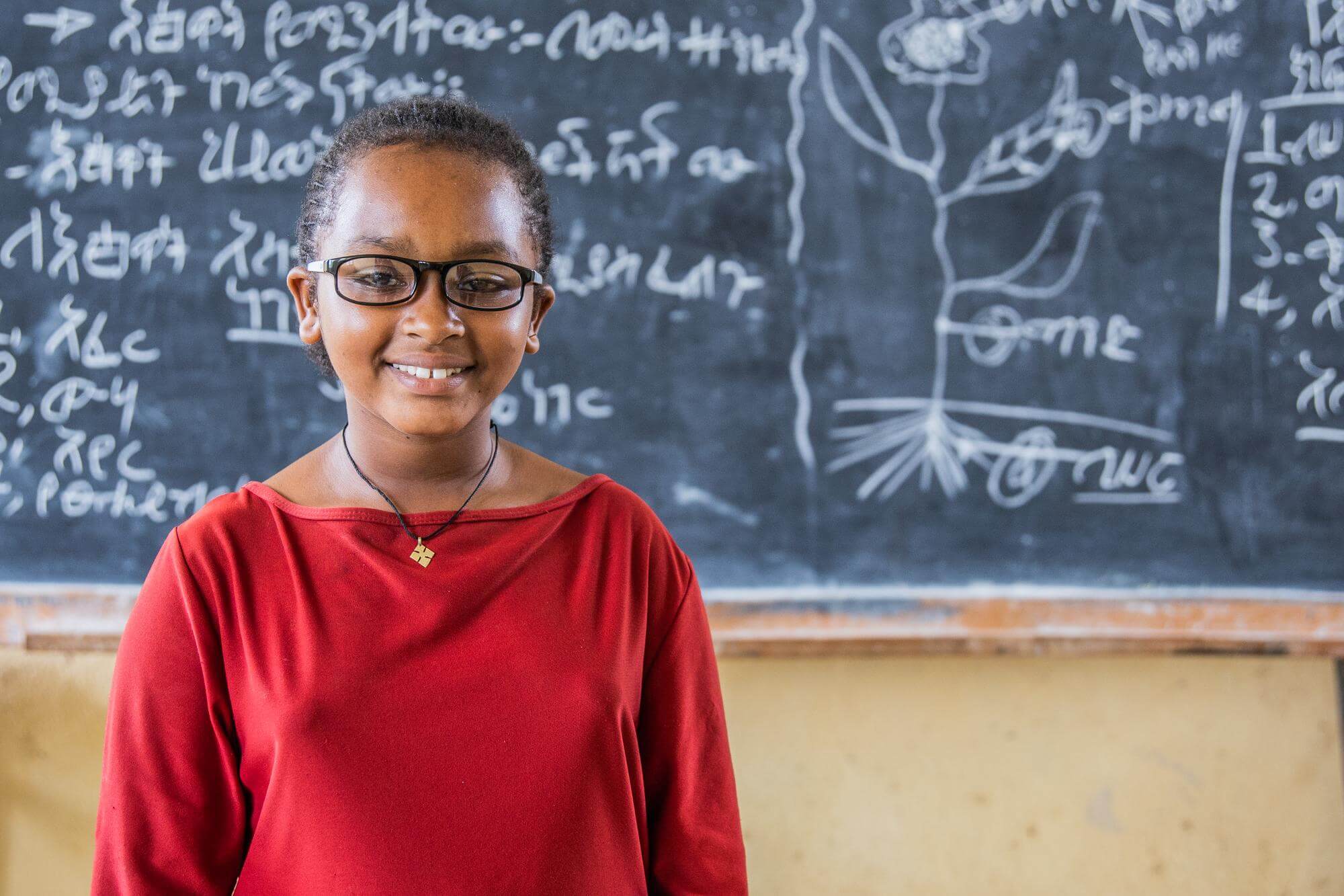 Image of Nigest Unche, standing and smiling in front of a blackboard in a school classroom. Nigest attends Sikela Primary School in Arba Minch, Ethiopia. She received glasses through 1, 2, 3 I can see! a school child eye health programme of Light for the World. ©Genaye Eshetu