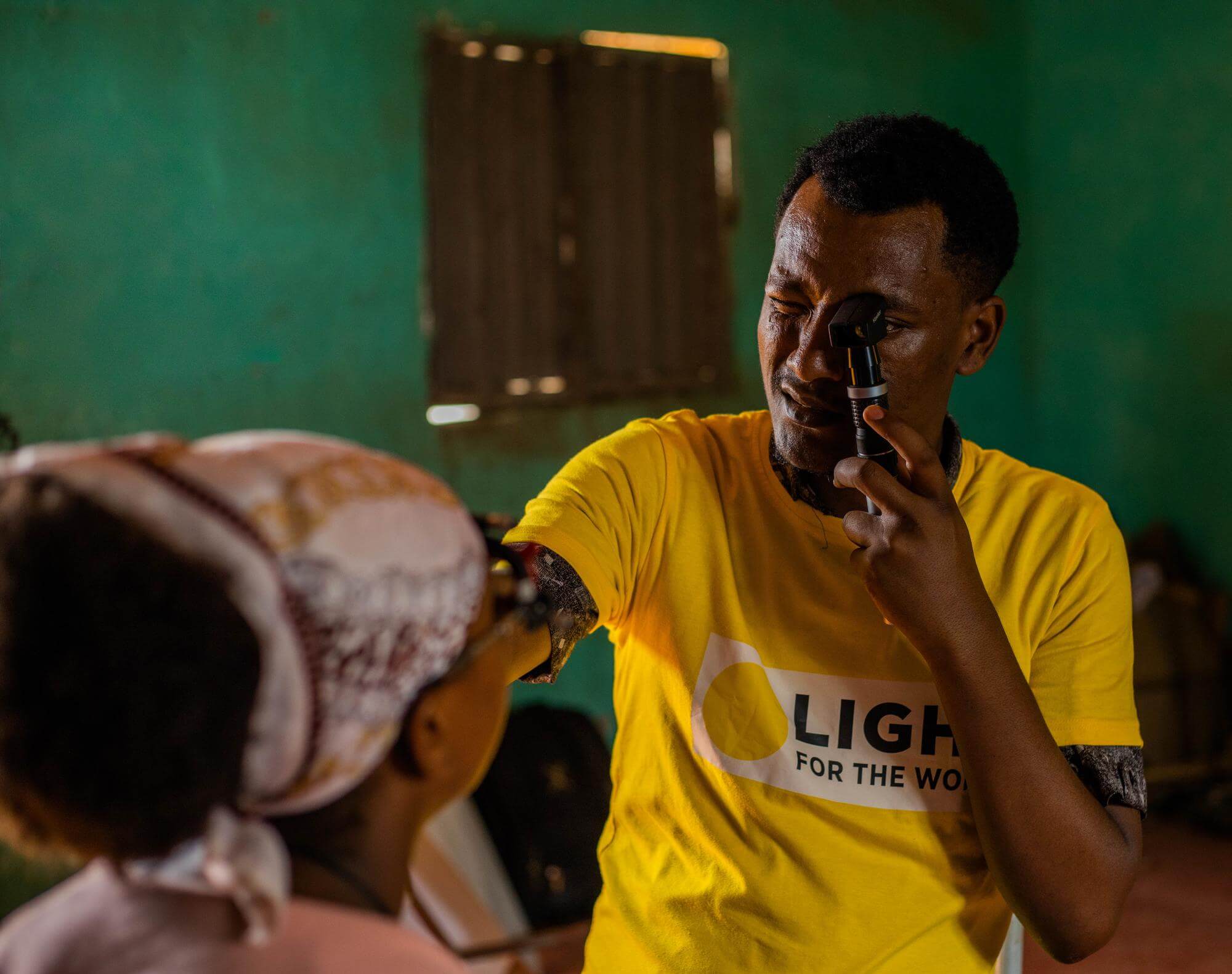 Image of schoolteacher Biruk Ayele, wearing a yellow Light for the World t-shirt, testing the eyes of pupil Senayet Bashura at Sikela Primary School in Arba Minch, Ethiopia.  The 1, 2, 3 I can see! programme, which focuses on child eye health, includes raising awareness with schoolteachers on how to detect potential eye problems in pupils. ©Lidya Alemayehu/Light for the World.