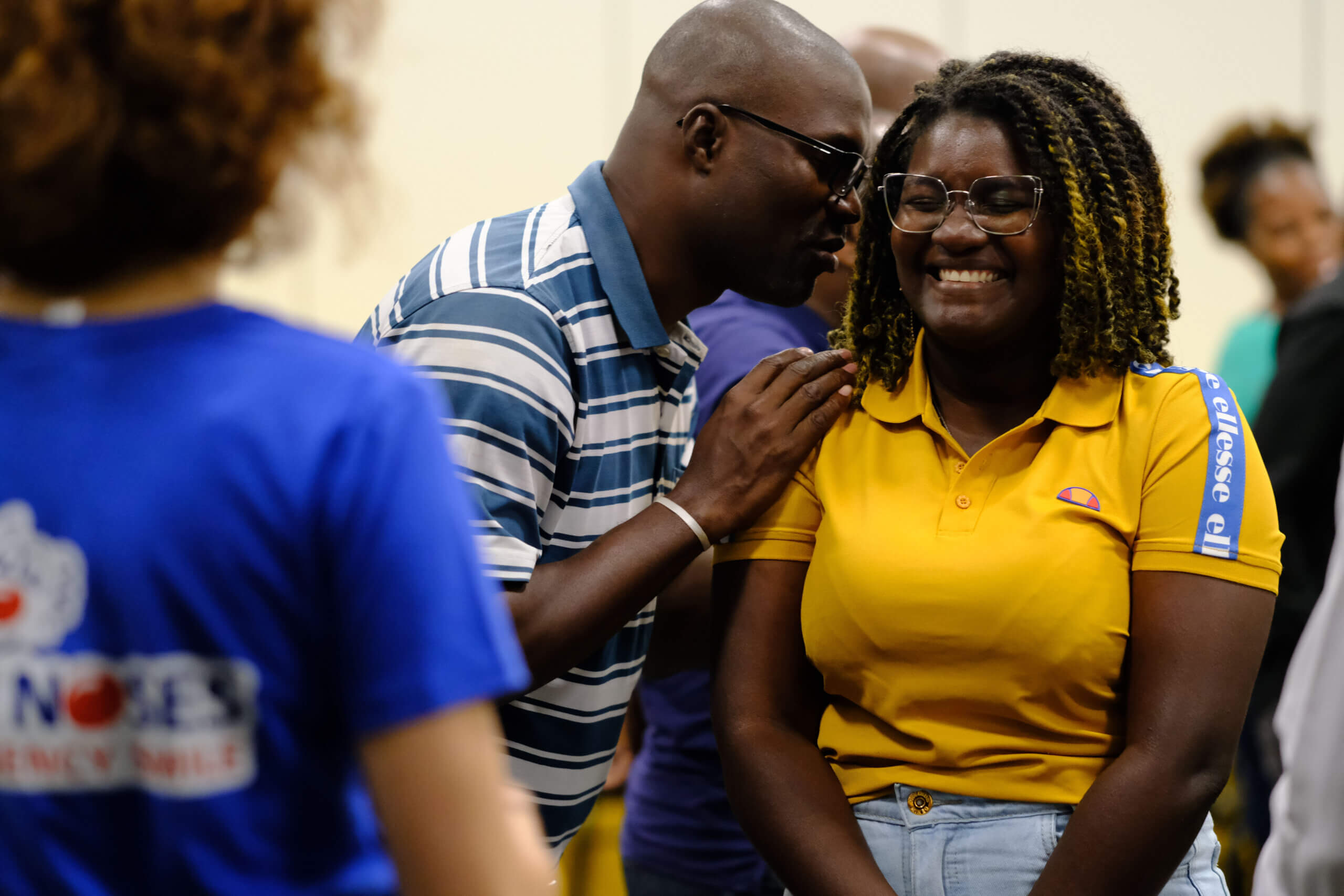 A woman is pictured laughing, wearing a yellow polo shirt, while a man wearing glasses and a blue and white striped top whispers into her ear.