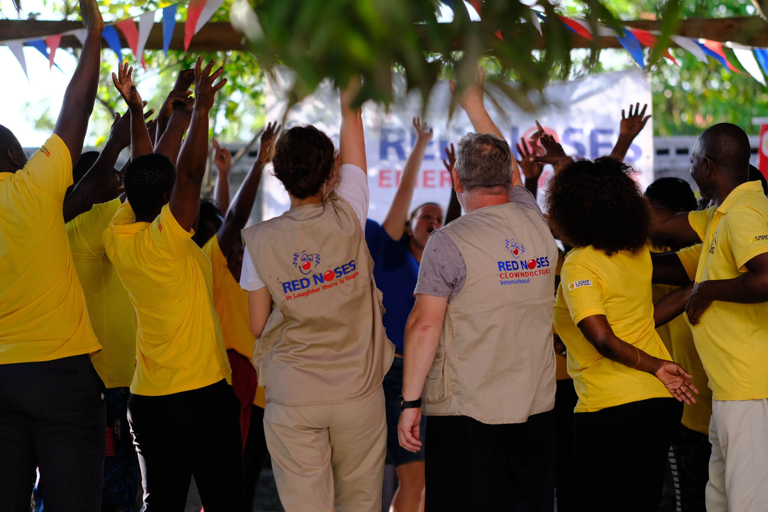 RED NOSES staff, wearing beige RED NOSES uniforms, Light for the World and OREBACOM staff raise their hands together during a Humour Relief Workshop. Breaking down stereotypes together in a celebratory mood.