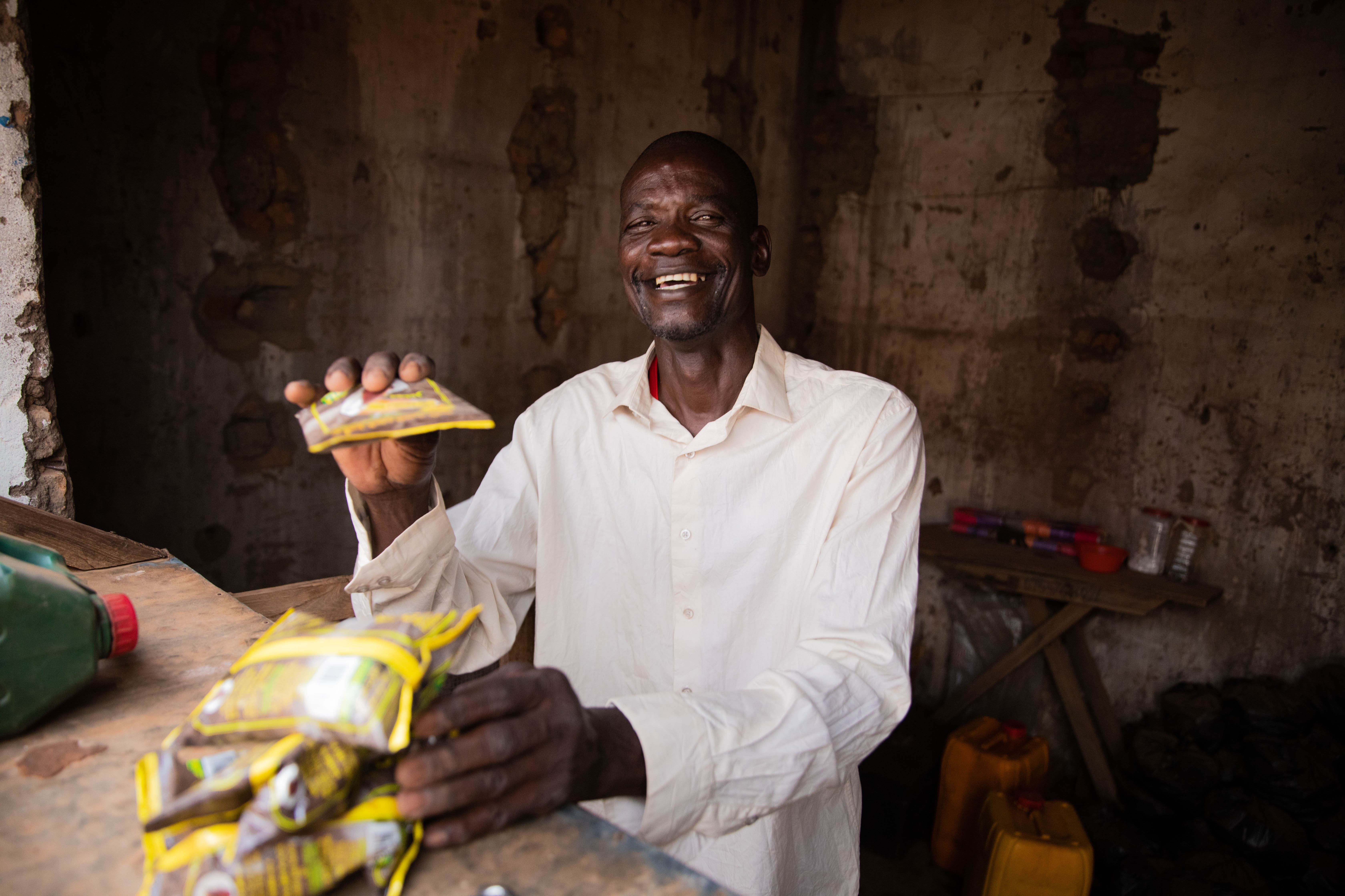 Celebrating OPDs on International Day of People with Disabilities. Peter smiles. He is wearing a pale pink shirt and is holding packets of food, which are brown and yellow. We can see some tools around him.