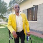 David Ndungu, disability inclusion field officer at Light for the World is wearing sunglasses, a bright yellow jacket, white shirt and black trousers. He is using crutches and holds a book.