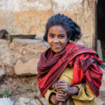 A young woman smiles as she looks into the camera. She is standing outside, in front of a brick wall, and wears a yellow dress, with a patterned red and maroon scarf around her neck. She is gently holding onto a wooden stick.