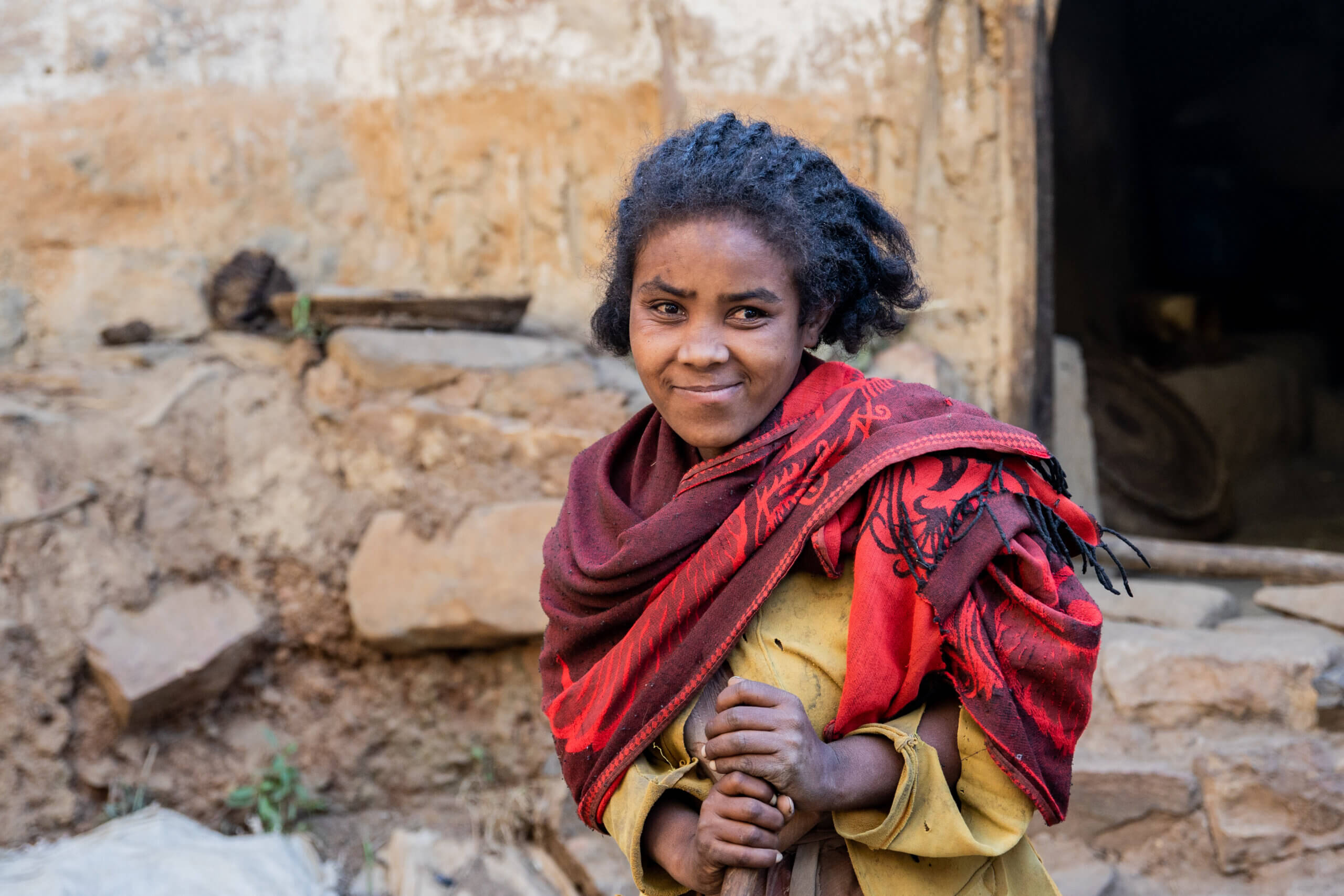 A young woman smiles as she looks into the camera. She is standing outside, in front of a brick wall, and wears a yellow dress, with a patterned red and maroon scarf around her neck. She is gently holding onto a wooden stick.