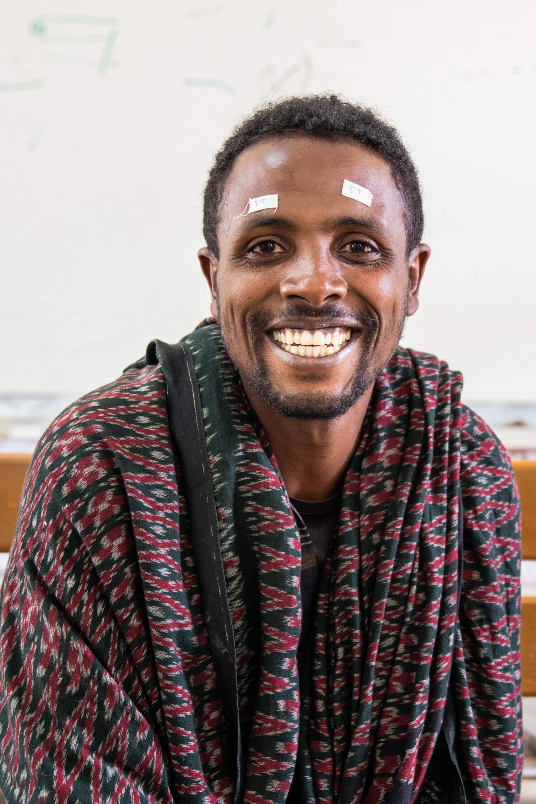 A man is smiling, with two small bandages on his head. He is wearing a red patterned jumper, with a t-shirt underneath.