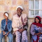 There are three people in the image, sitting in front of a wall. The boy is on the left, wearing a bandage over his eye, with his family on the right. They are all smiling.