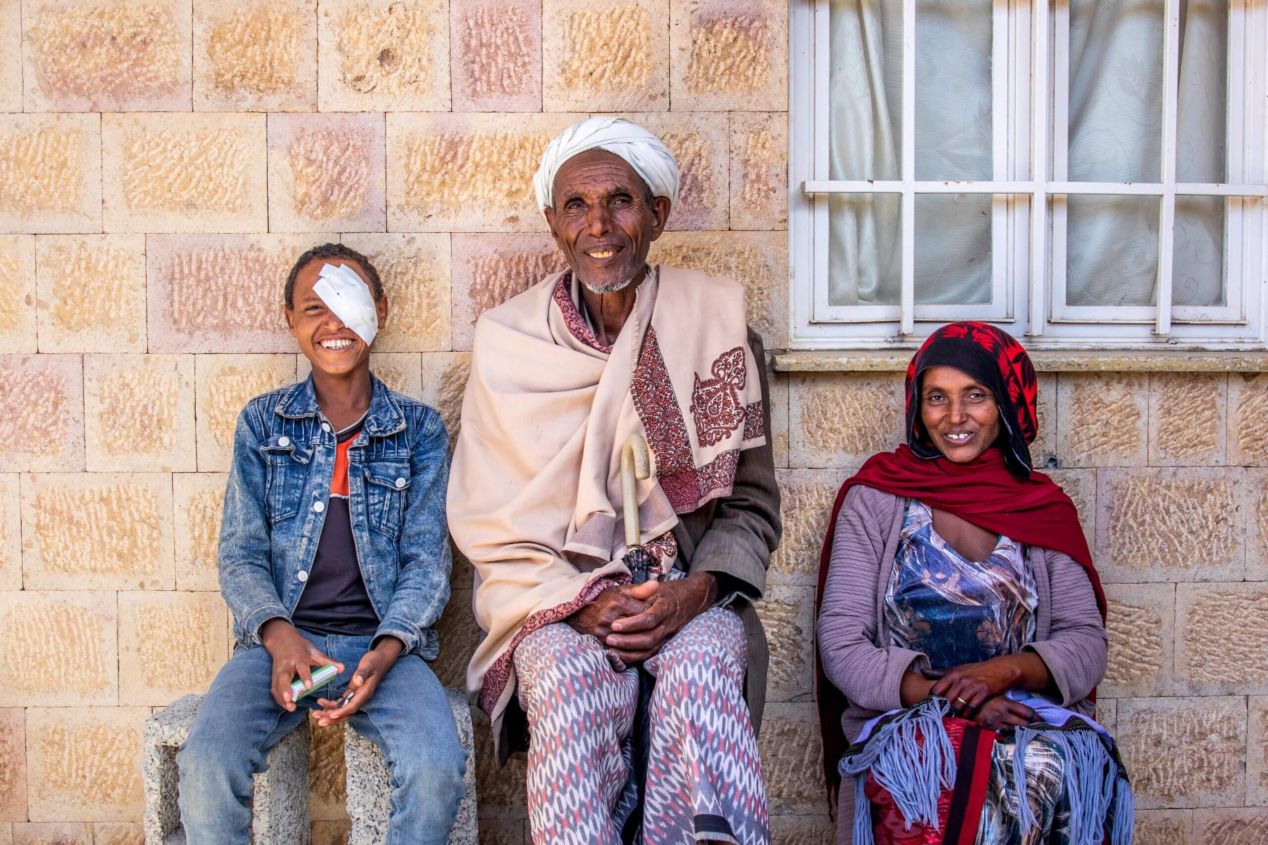 There are three people in the image, sitting in front of a wall. The boy is on the left, wearing a bandage over his eye, with his family on the right. They are all smiling.