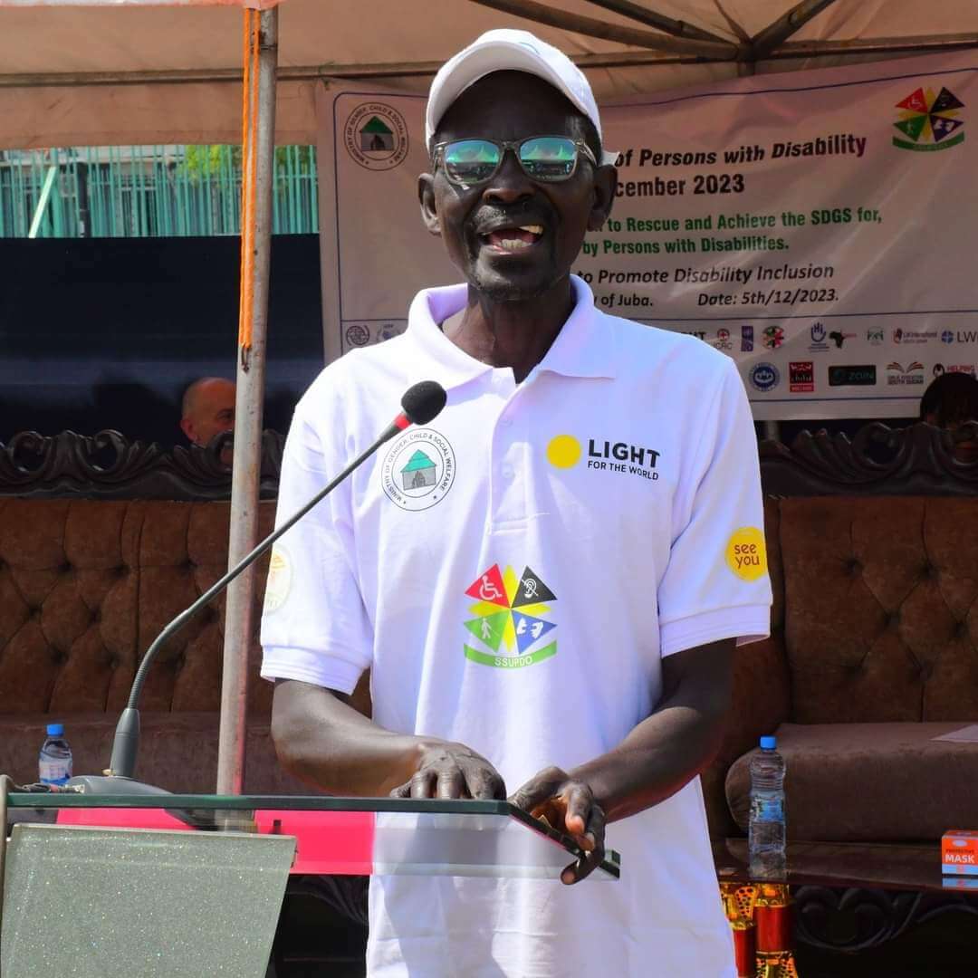 A man wearing glasses, a white hat and white tshirt stands at a podium in front of a microphone. His t-shirt has the logos of Light for the World, Ministry of Gender, Child and Social Welfare and SSUPD.