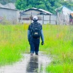 Image of Daniel Anyang, a Disability Inclusion Facilitator at Light for the World, visiting flooded communities in Jonglei state in South Sudan. Daniel has his back to the camera and is walking towards homes. He is walking through a flooded area of grassland. The water is just below his knees.