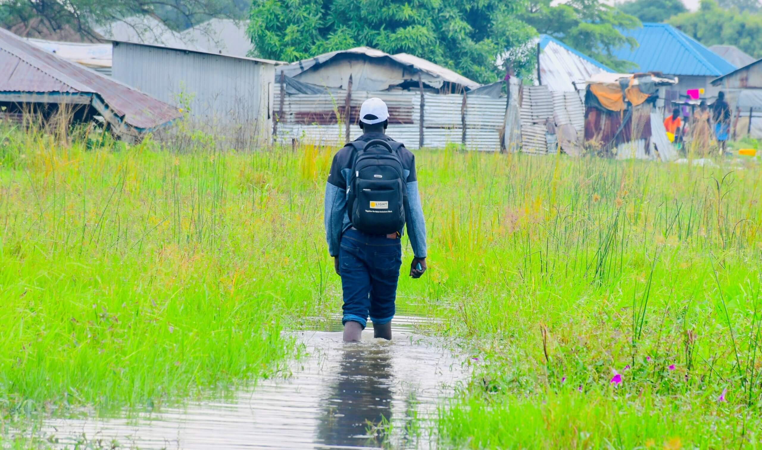 Image of Daniel Anyang, a Disability Inclusion Facilitator at Light for the World, visiting flooded communities in Jonglei state in South Sudan. Daniel has his back to the camera and is walking towards homes. He is walking through a flooded area of grassland. The water is just below his knees.