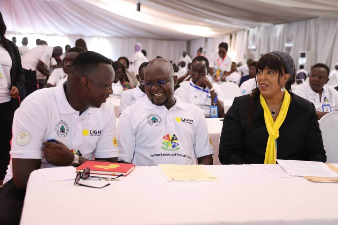 Two men sit on the left next to a woman in a black jacket and bright yellow scarf. The men are wearing Light for the World t-shirts