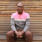 Image of Stanley Mutuma, a renowned disability rights activist and lawyer who has joined Light for the World’s international board. Stanley is wearing a pink and grey top and sat on a wooden bench.