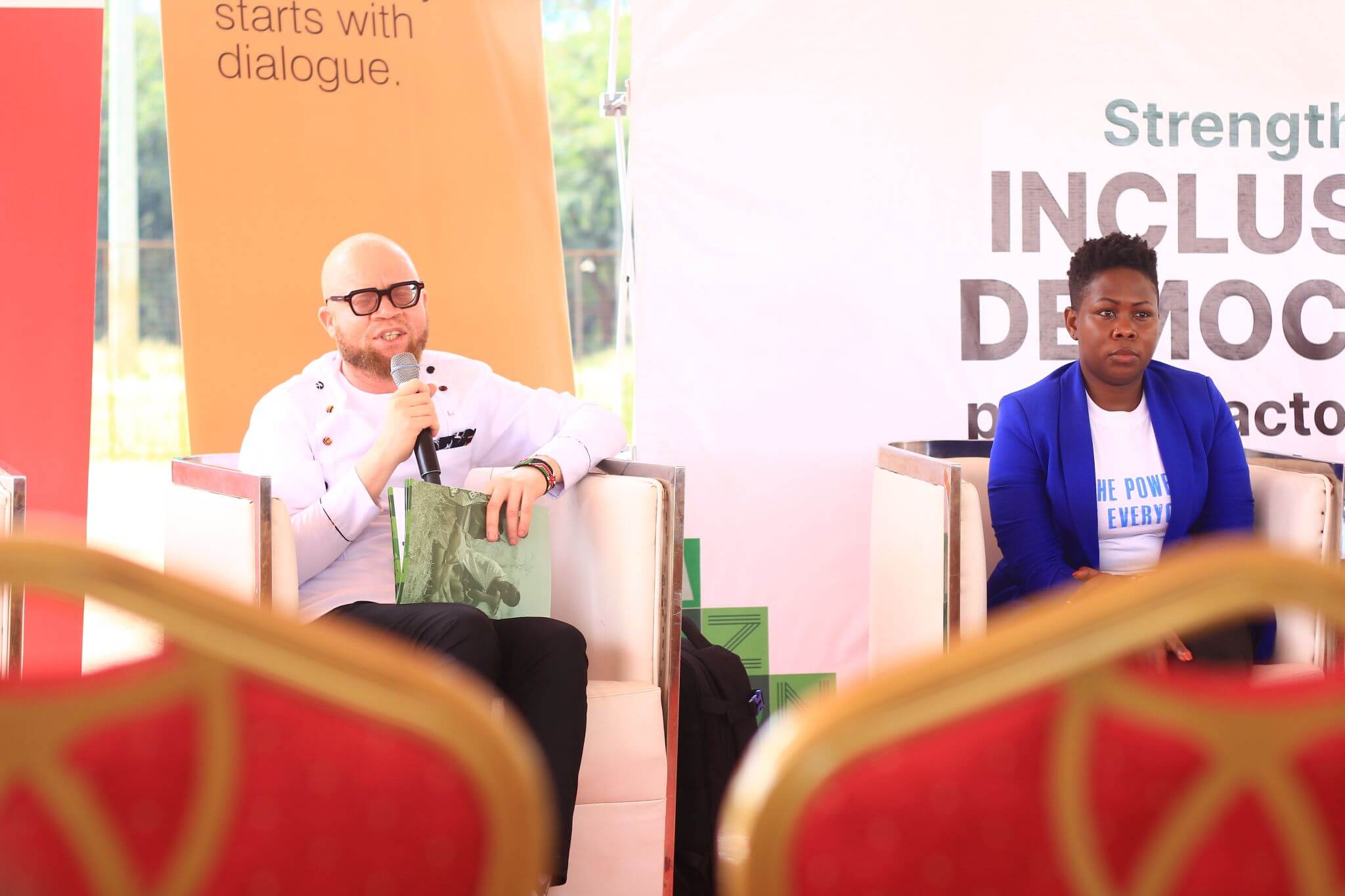 Collins Ombajo, sits on the left hand side of the picture in a white leather chair. He is wearing black rimmed glasses and speaks into a microphone. A woman sits to the right wearing a white t-shirt and bright blue jacket. They are sitting in front of big red, orange and white posters. 