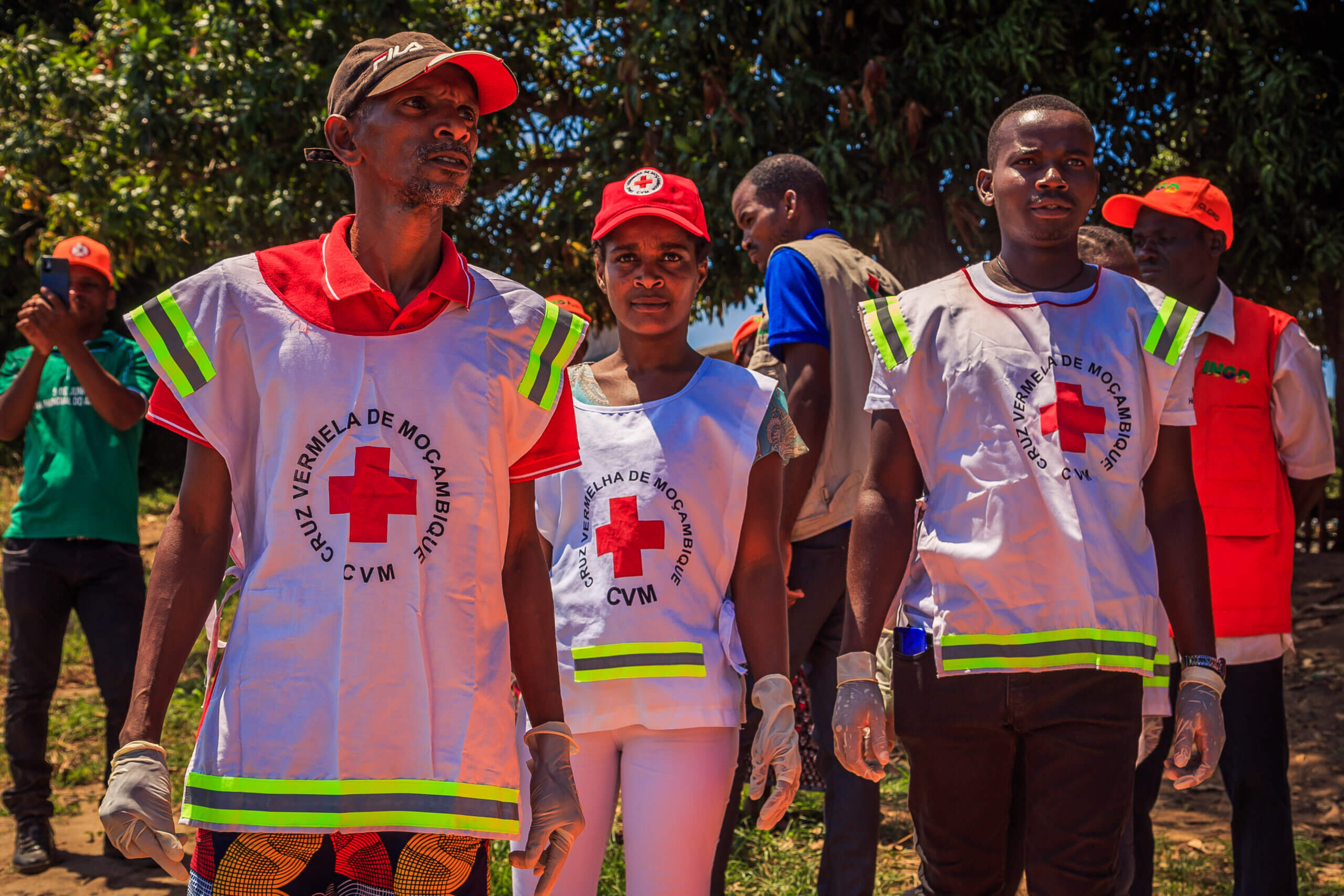A woman stands in the middle of two men, wearing a red cap. All three people are wearing white vests, with reflective stripes and crosses the middle. The vests read "CVM. Cruz Vermela De Mocambique". 