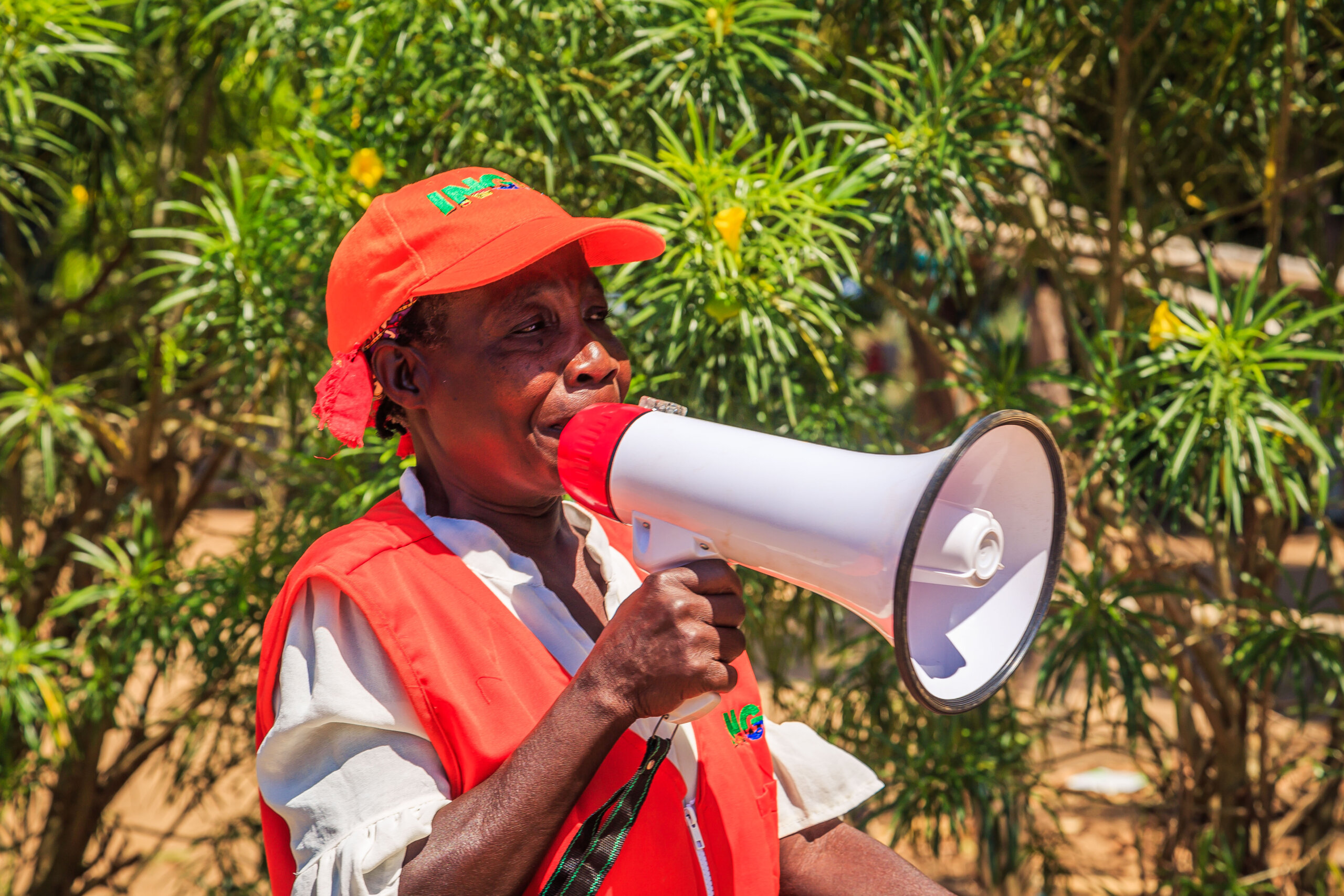A woman takes part in a Cyclone Simulation exercise in Buzi, Mozambique as part of the ICDP project, funded by Austrian Development Agency. Gender is mainstreamed throughout the project, ensuring that women and girls with disabilities are included in creating emergency response plans to climate-related disasters. She is wearing an bright orange hat and vest, and is using a megaphone. She's standing outside in front of some vegetation.