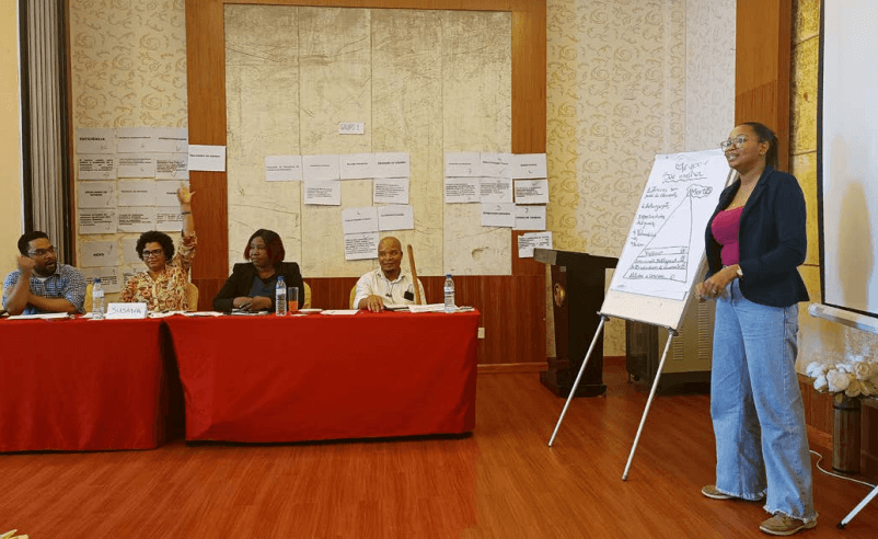 Five people participate in a workshop. One woman stands next to a flipchart, while four others are seated at a red desk. There are lots of sheets of paper pinned to the wall.