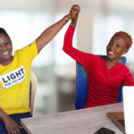 Two women sitting at a desk holding up their hand together, celebrating partnership. The woman on the left wears a yellow Light for the World t-shirt, the woman on the right wears a red blouse.