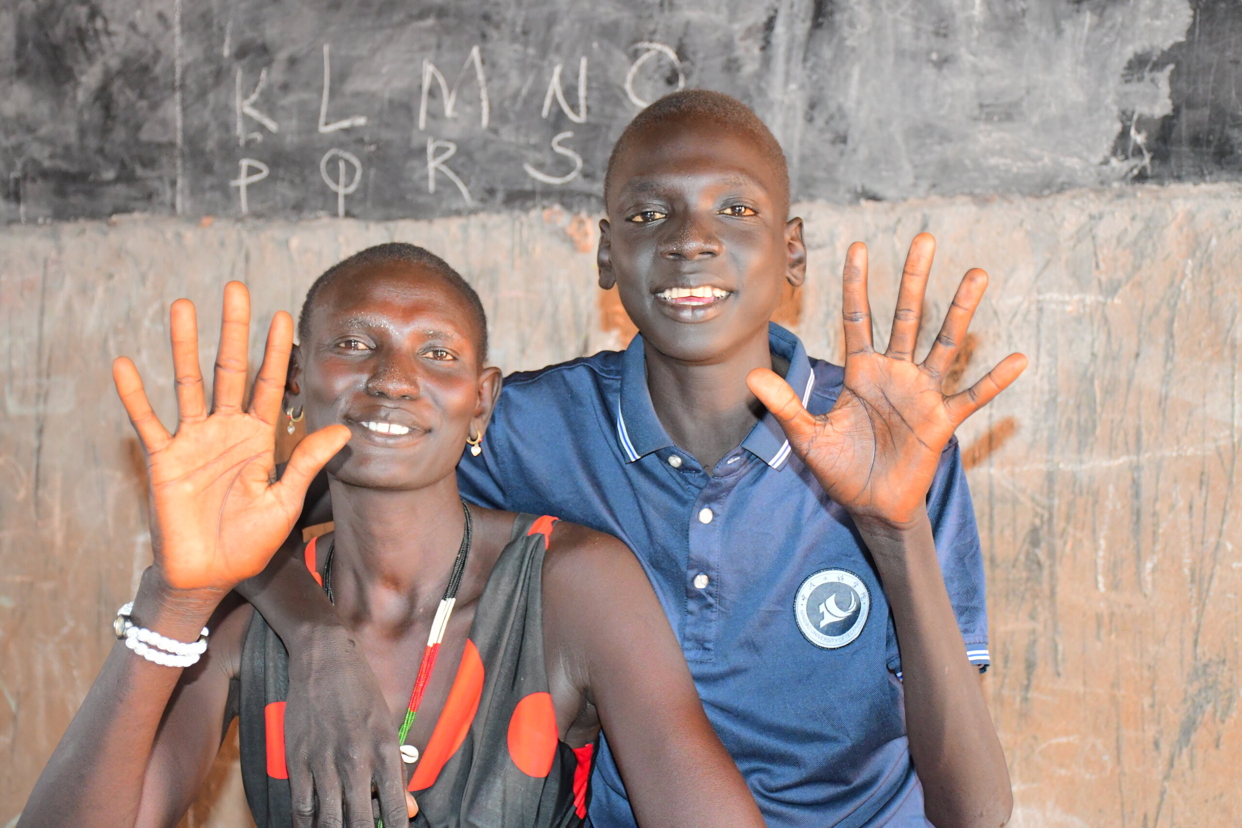 Two people smile in front of a blackboard, holding their hands spread out, with palms to the camera. A boy sits on the rights, wearing a blue polo-shirt, he has his arm around his mum's shoulders - to the left - who is wearing a black and orange polka dot top and a white pearl bracelet.