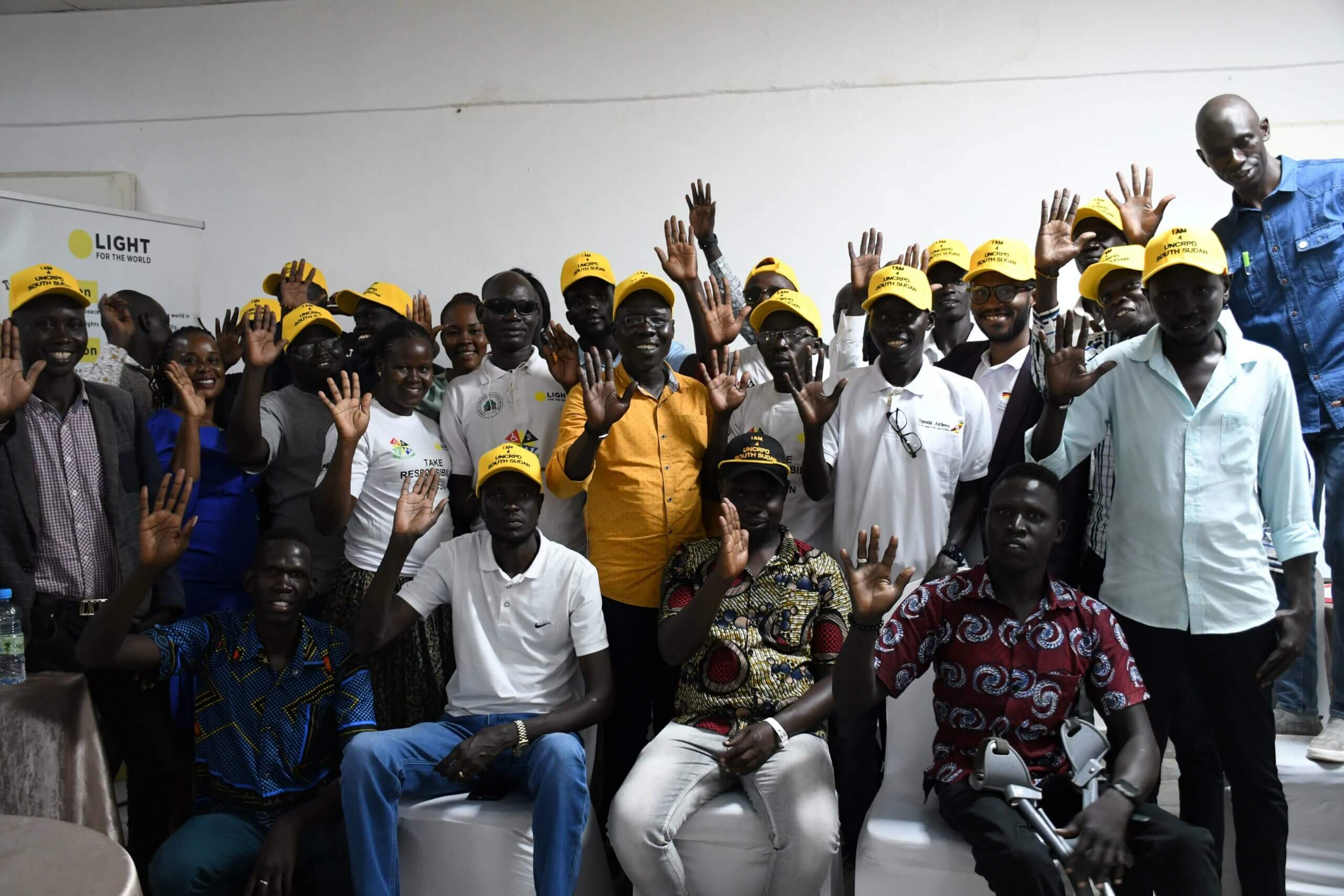 Journalists from South Sudan attend a training session on disability inclusive language by Light for the World. There are about 20 journalists, most are smiling at the camera and waving. 