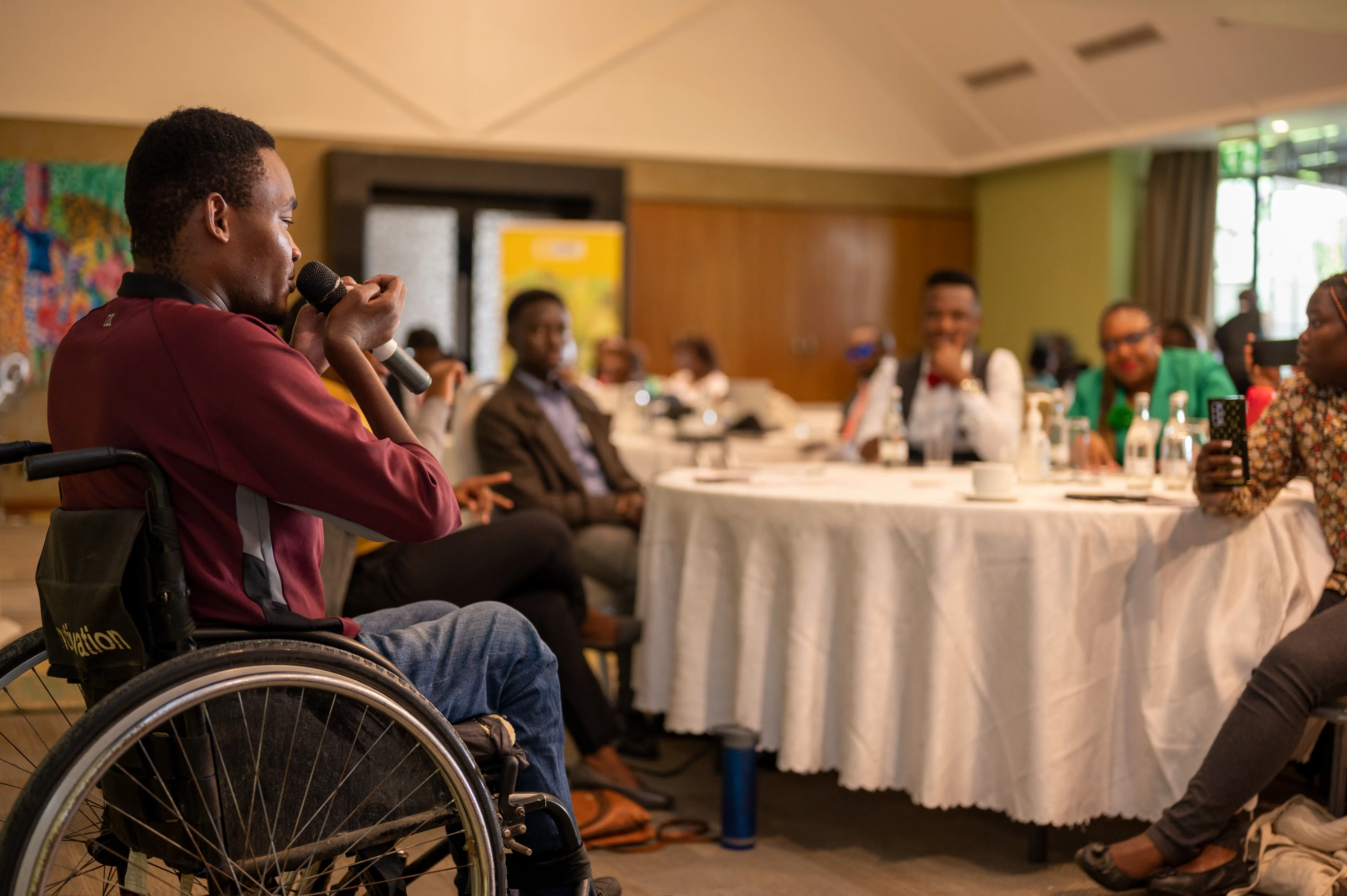 A man in a wheelchair is holding a microphone and speaking to an audience of people seated around tables at the We Can Work Inception Meeting.