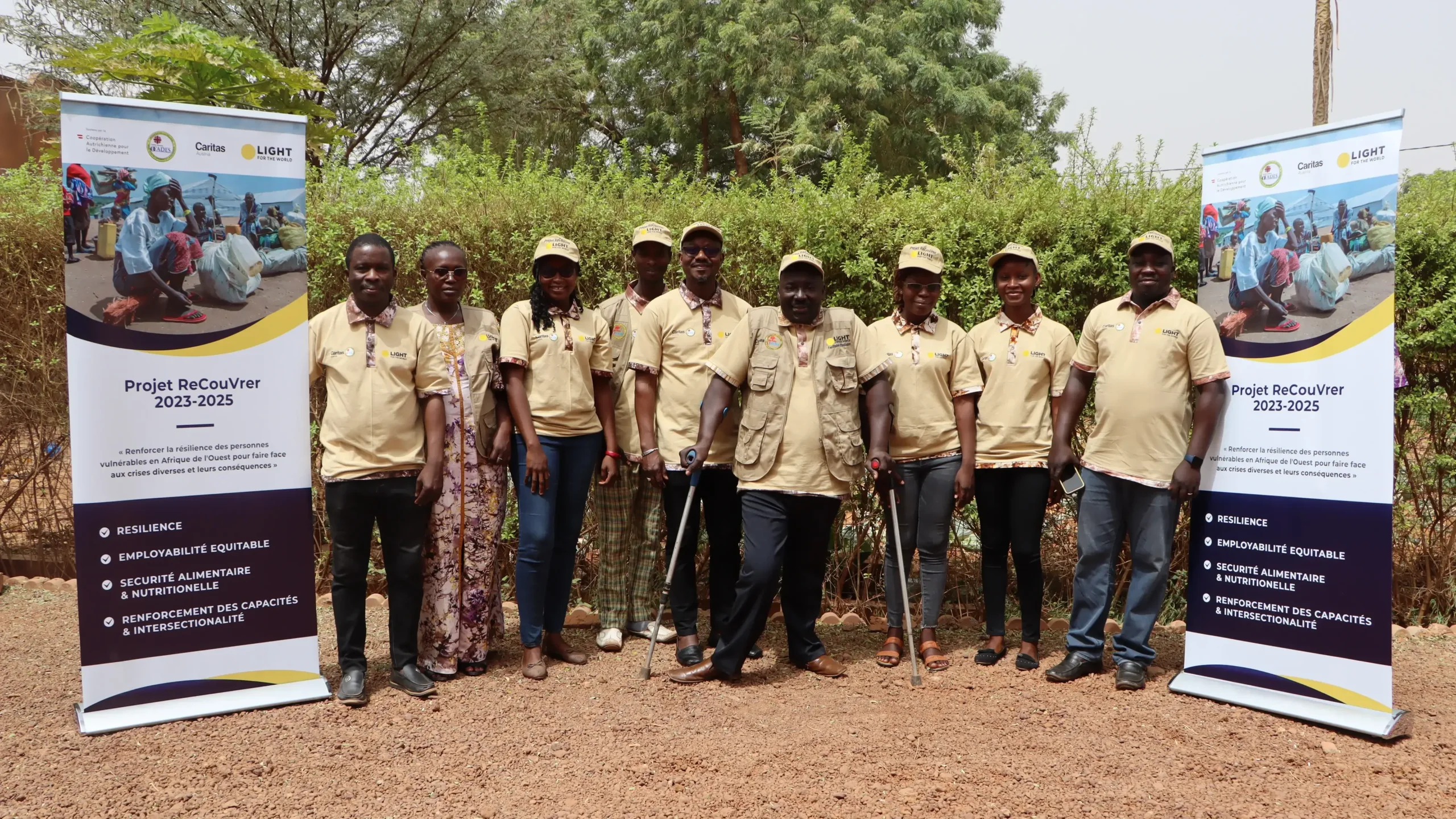 A group of people wearing beige Light for the World clothing and caps is posing for a group photo. The man in the middle is holding clutches. To both sides of the group stand info panes, giving information about the Recouvrer project.