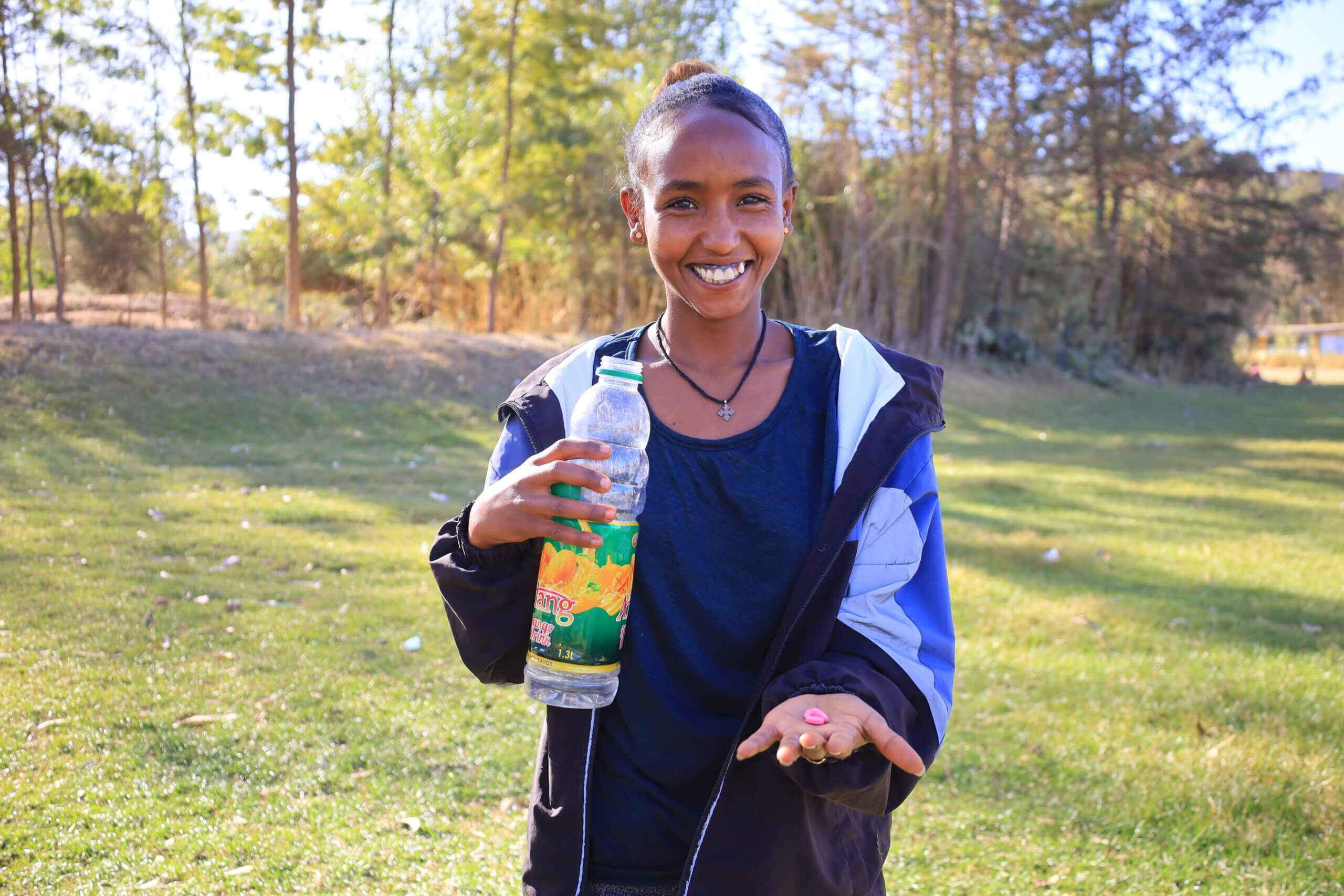 A girl smiles, standing outside on grass. It is sunny and there are trees behind her. She holds a water bottle in one hand and a small pill in the other.