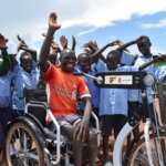 Moses on his new tricycle surrounded by his classmates. Moses has his arm raised and is smiling. His classmates are also smiling.