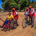 People take part in a Cyclone simulation exercise in Buzi district. Helena Rego, a DIF who uses a wheelchair, is supported by two people in red high-visibility jackets and hats. Three other men are also in high-vis clothing. One has a megaphone.