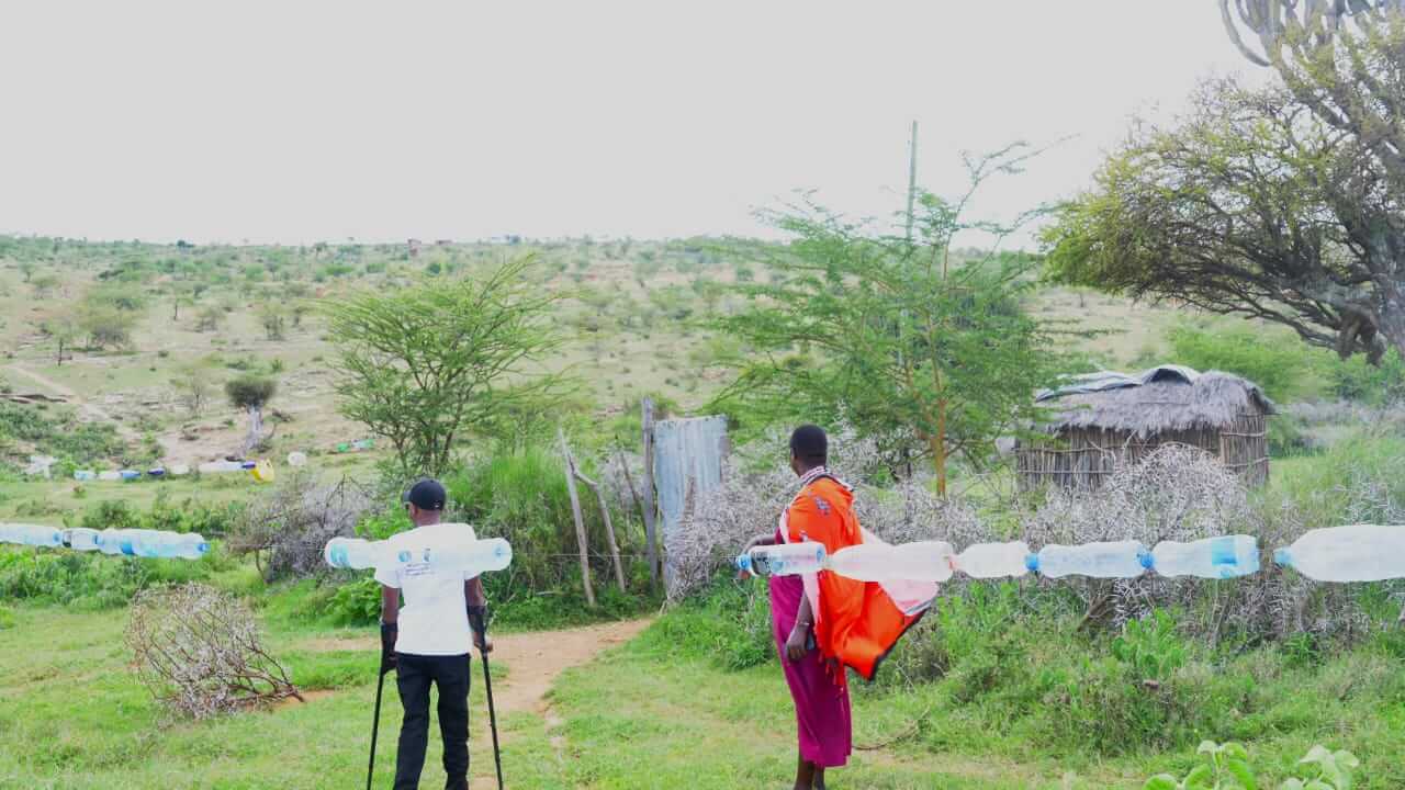 Two people stand in a field looking into the distance at green. There is a line of plastic bottles closer to the camera than the people. One woman is wearing an orange and red outfit, and David Ndungu is on the left in a white top and black trousers, using crutches.