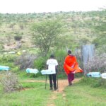 Two people stand in a field looking into the distance at green. There is a line of plastic bottles closer to the camera than the people. One woman is wearing an orange and red outfit, and David Ndungu is on the left in a white top and black trousers, using crutches.