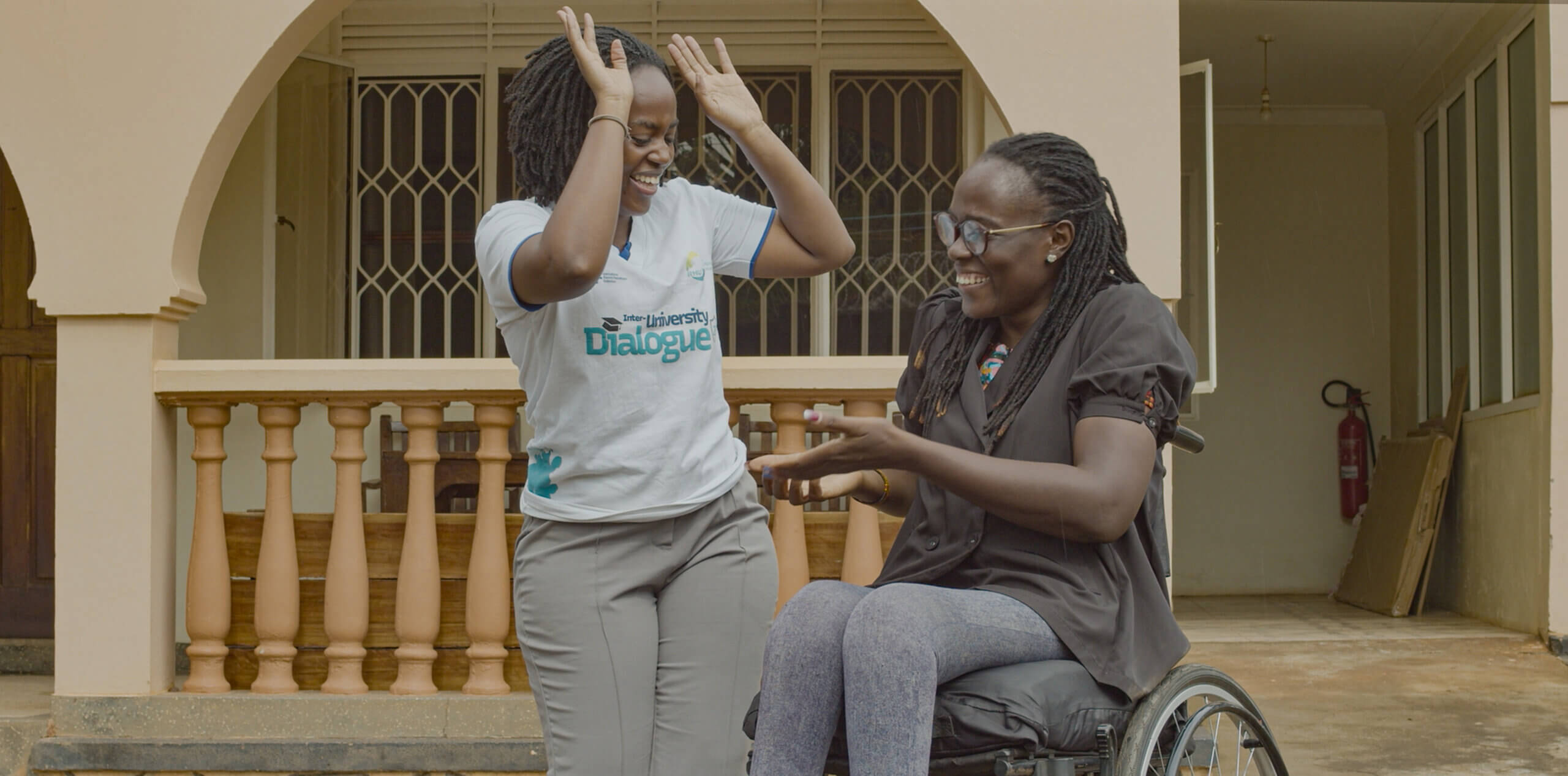 Two women are outside smiling and high fiving with all hands. Doreck stands up wearing a white tshirt and Naome sits in a wheelchair with a grey shirt and leggings. You can tell they are good friends and having fun together,