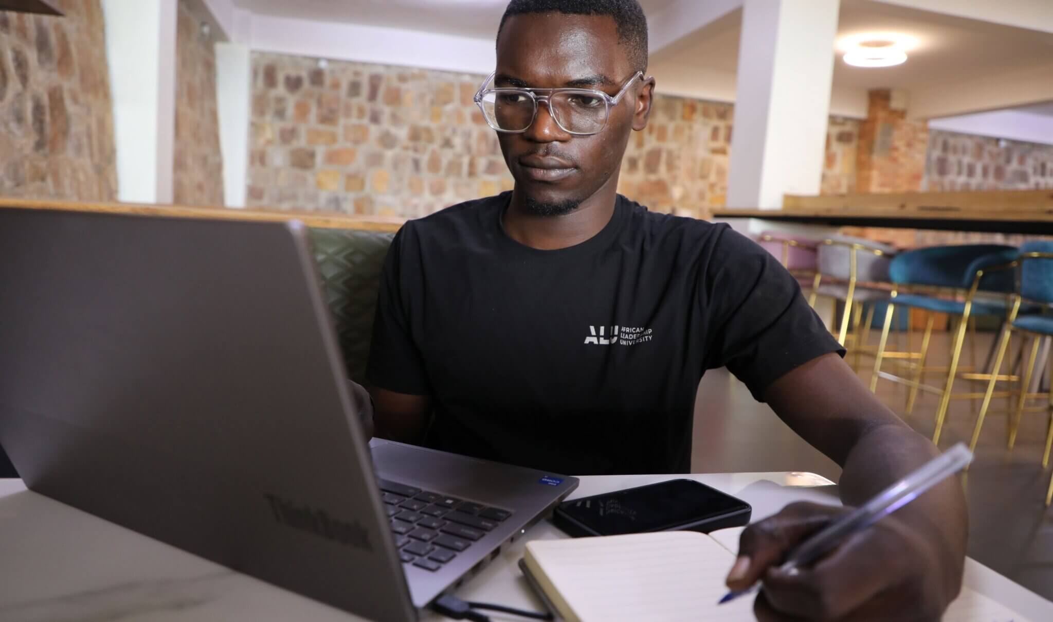 Patrick Mfitumkiza, one of the winners from Rwanda. Patrick is looking at a laptop and taking notes with a pen and pad.