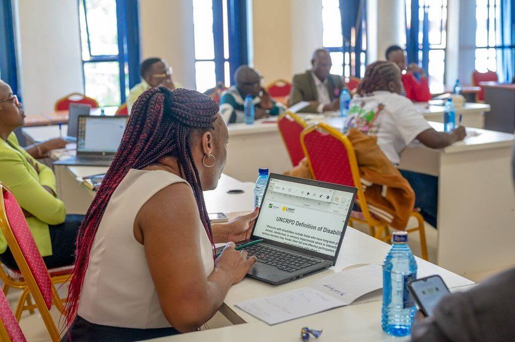 A group of people take part in a workshop. A woman is closest to the camera. She wears a white top and long red and black braids. She is sitting at a white desk, on a red and gold chair, looking at a laptop, which reads "UNCRIPD Definition of Disability....". There are several people in the backround, all at the same desks and chairs, with water bottles and paper around. They are all looking towards the front of the room. It is out of view but seems like someone is delivering a presentation.