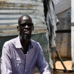 A man smiles. He is sitting outside a corrugated iron shelter. He wears black sunglasses and a purple striped shirt. He has a beard. It looks hot outside and you can see sun shining in the background.
