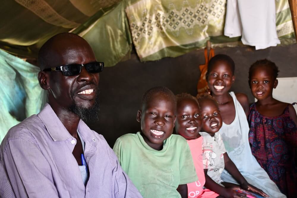 A group of people smile for a picture. They are inside a shelter with a green patterned fabric roof. A man sits at the front wearing sunglasses, a purple shirt and white vest. Next to him are a group of 5 children, all with big smiles on their faces. The three closes to David sit next to him, while the two children in the back stand.