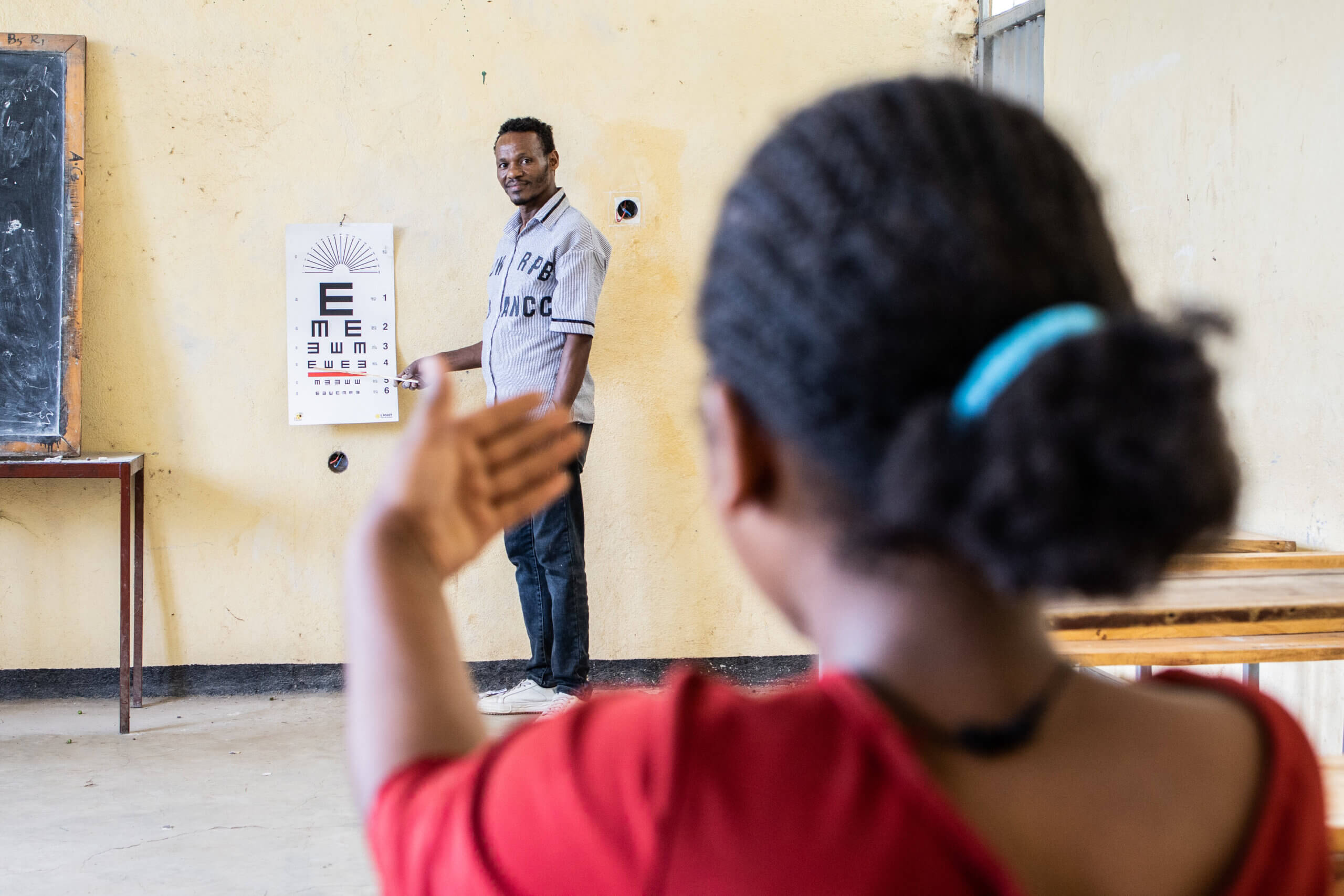Inside a classroom a man stands in front of a beige wall, with an eye test attached to the wall. There are different letters and shapes. A girl is close to the camera, with her back to it. She has one hand up, wears a red tshirt and her hair is tied in a blue headband.