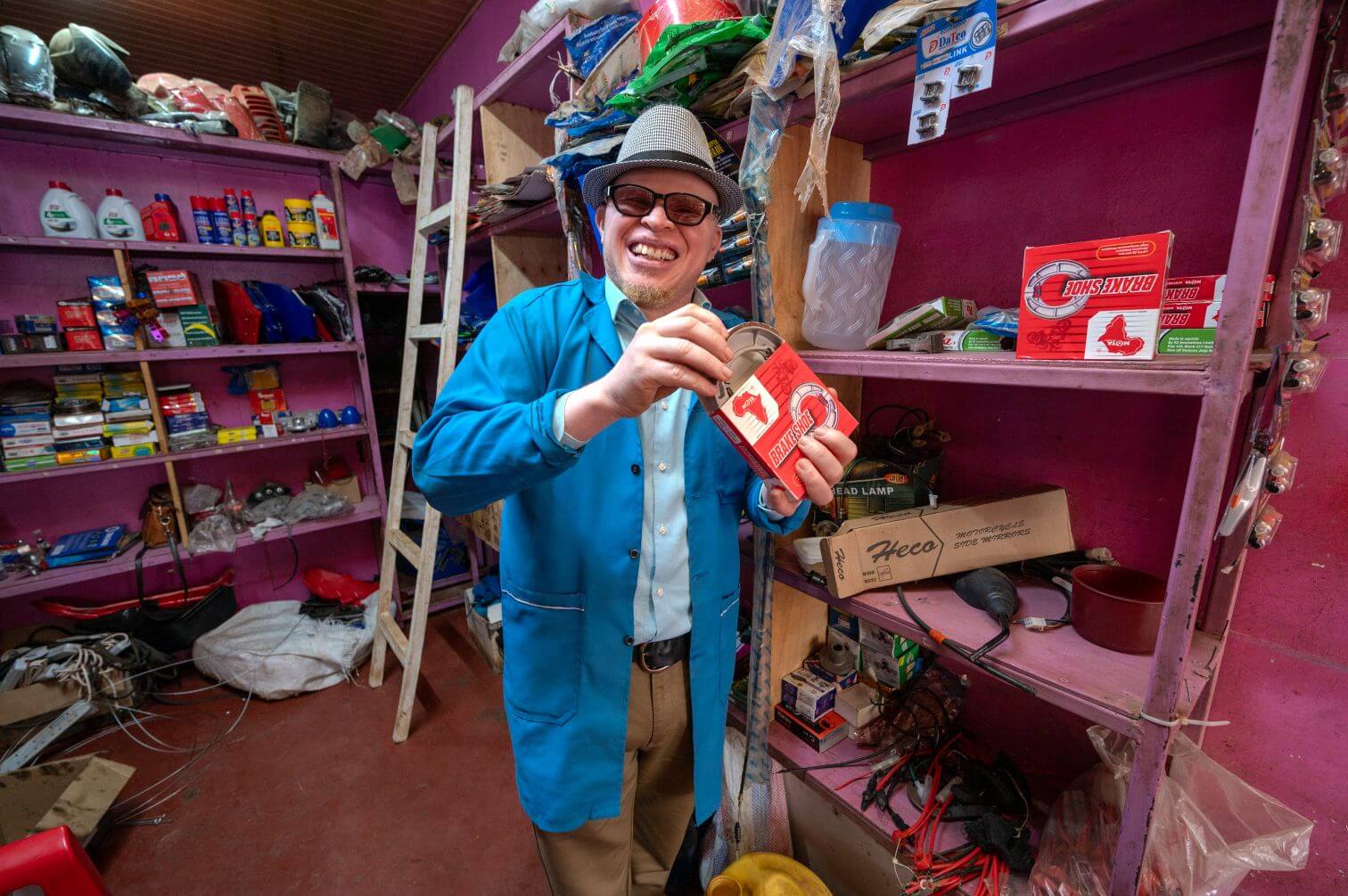 Anthony Maina in the storeroom at his garage. The storeroom is painted pink, with various tools and equipment for motorcycle repair on the shelves. Anthony is dressed in a blue coat and wearing a hat. He is holding a Brake Shoe part and smiling at the camera.