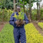 Duncan Mwirichia, a landscape gardener and entrepreneur who has a hearing disability. Duncan is a Kenyan man, wearing blue overalls. He is holding a green plant and standing in front of a row of plants. He is smiling.