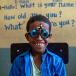 Kedir Mohamed, a young Ethiopian boy sits smiling in a classroom while wearing a blue eye-testing frame used for vision screening. He wears a bright blue shirt over a pale green T-shirt. Behind him, the wall is painted yellow and blue, with hand-painted text in English that reads questions like “What is your name?” and “How old are you?” The boy appears happy and engaged during a school-based eye health screening.