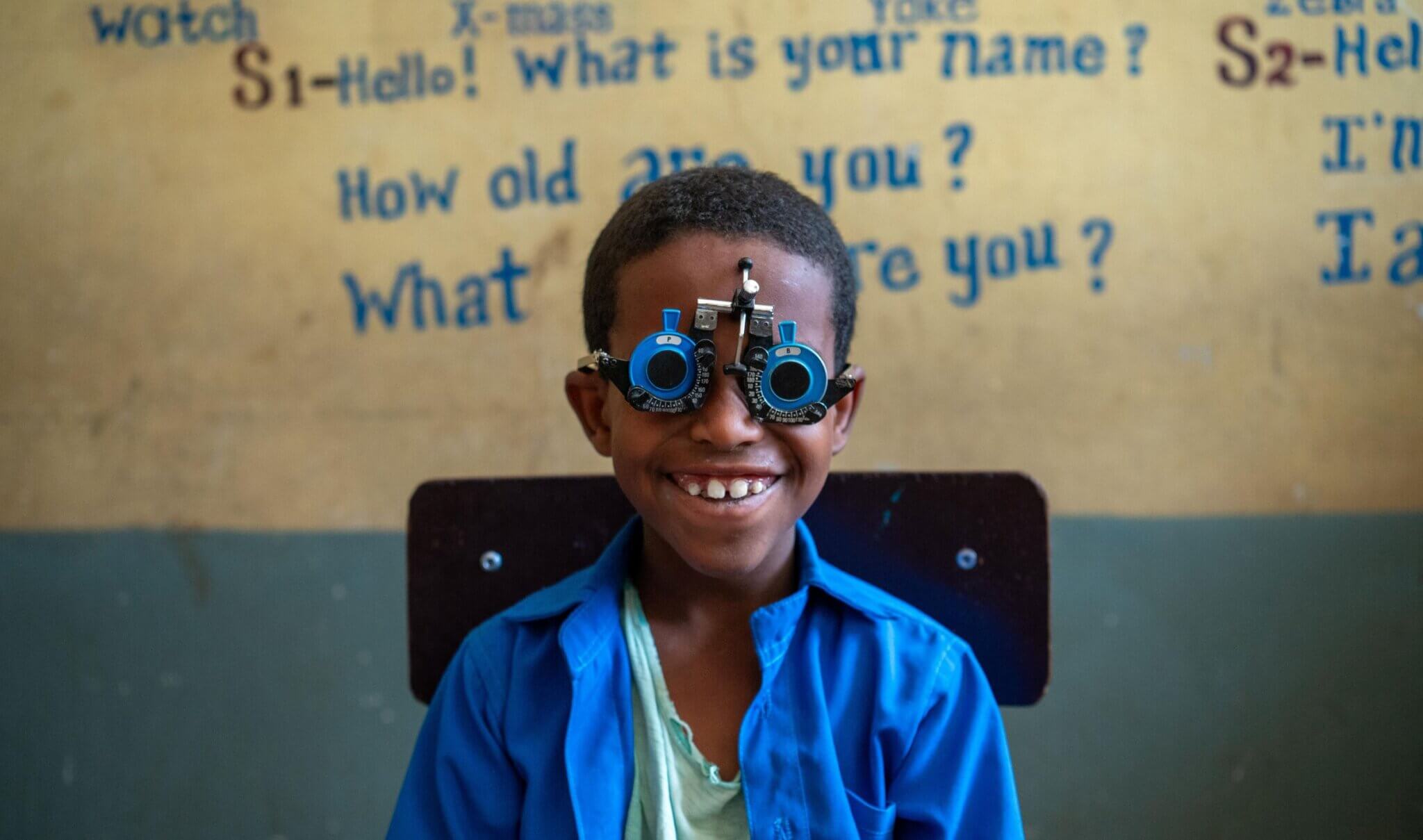 Kedir Mohamed, a young Ethiopian boy sits smiling in a classroom while wearing a blue eye-testing frame used for vision screening. He wears a bright blue shirt over a pale green T-shirt. Behind him, the wall is painted yellow and blue, with hand-painted text in English that reads questions like “What is your name?” and “How old are you?” The boy appears happy and engaged during a school-based eye health screening.