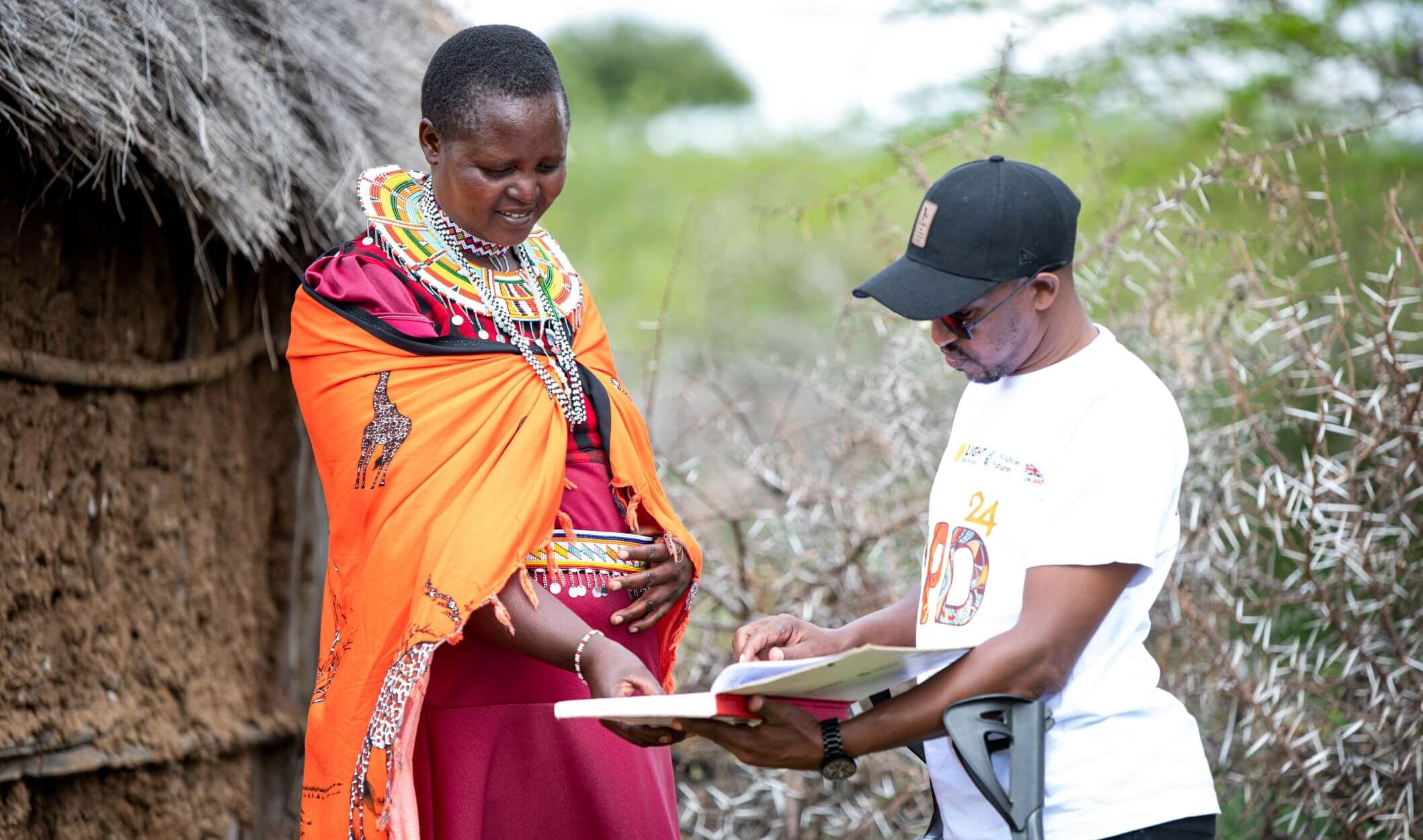 Kushuke Legei with David Ndungu, Disability Inclusion Field Officer at Light for the World. Kushuke is wearing an organge shawl with colourful clothes and jewellery. She is looking at a training manual with David, who points something out on a page. They are in Kushuke's home village. 